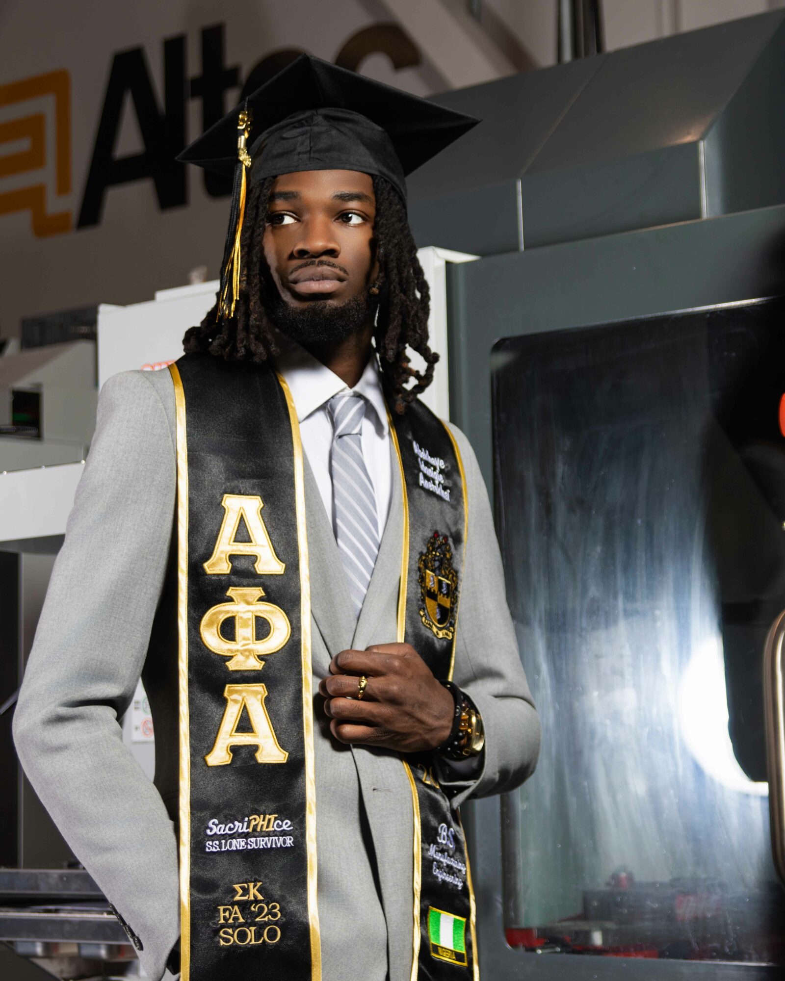 Graduate in cap and gown with honor cords, standing near a machine, holding a diploma, indoors. Word 'AΦA' visible.