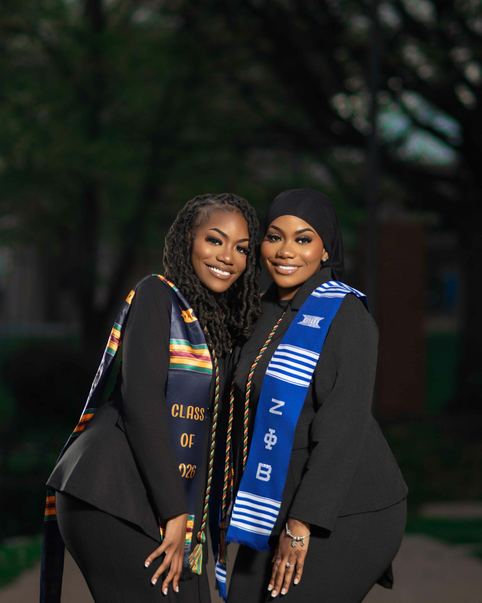 Two women in graduation gowns and stoles smiling outdoors, with trees in the background.