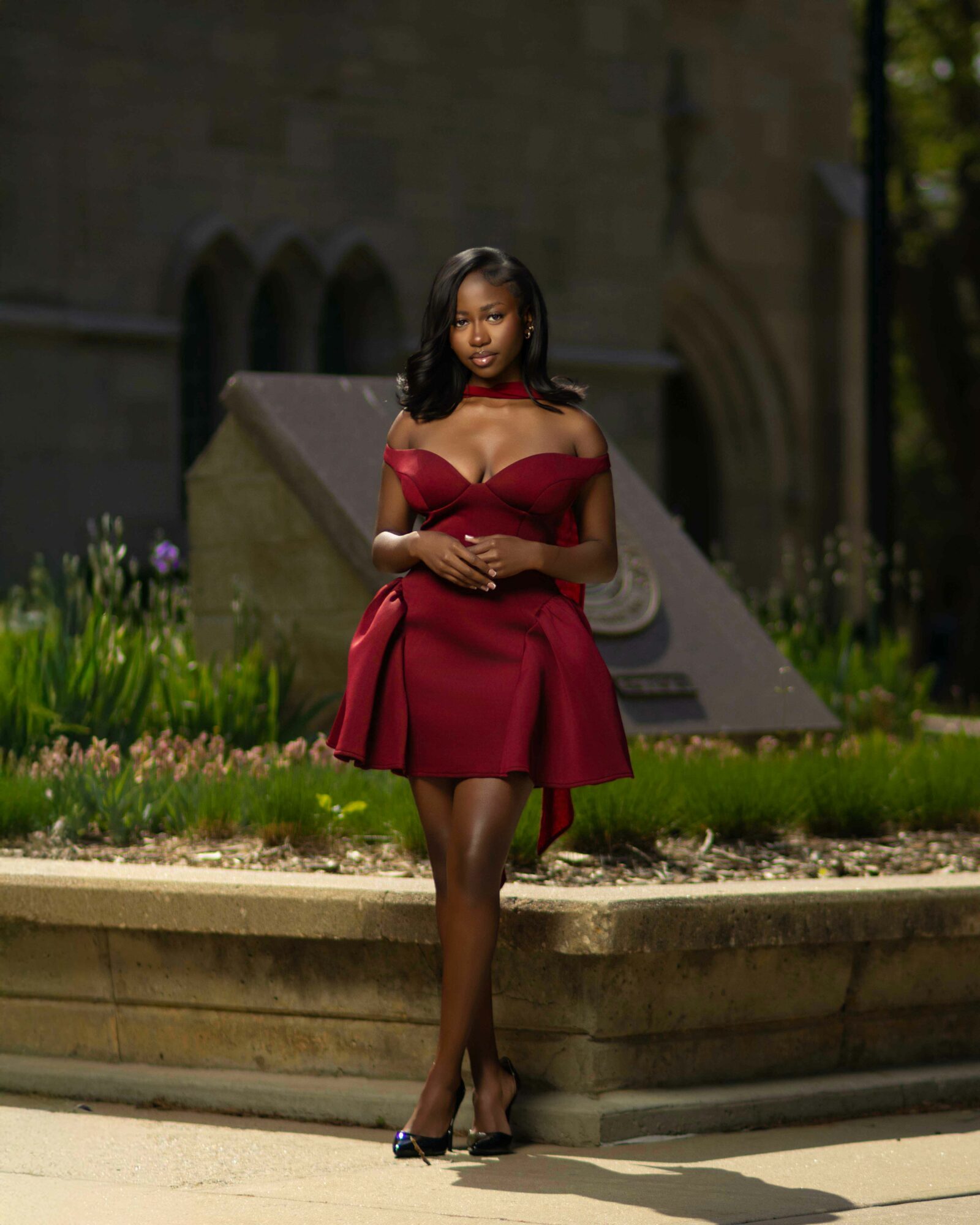 Woman in red dress standing outdoors near a stone monument, with greenery and flowers around her.