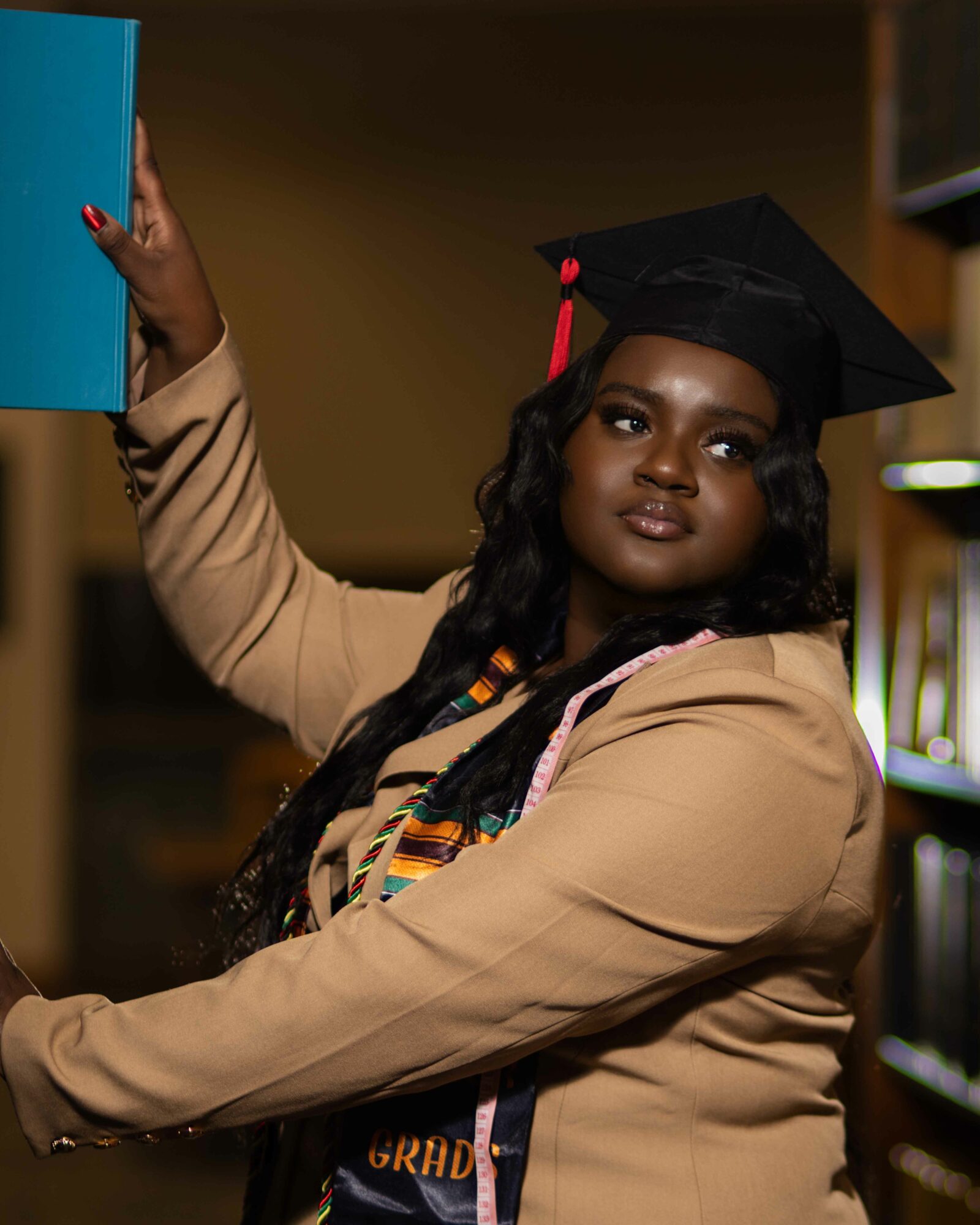 Graduate woman in cap and gown taking a selfie in a library, holding a blue book, with bookshelves in background.