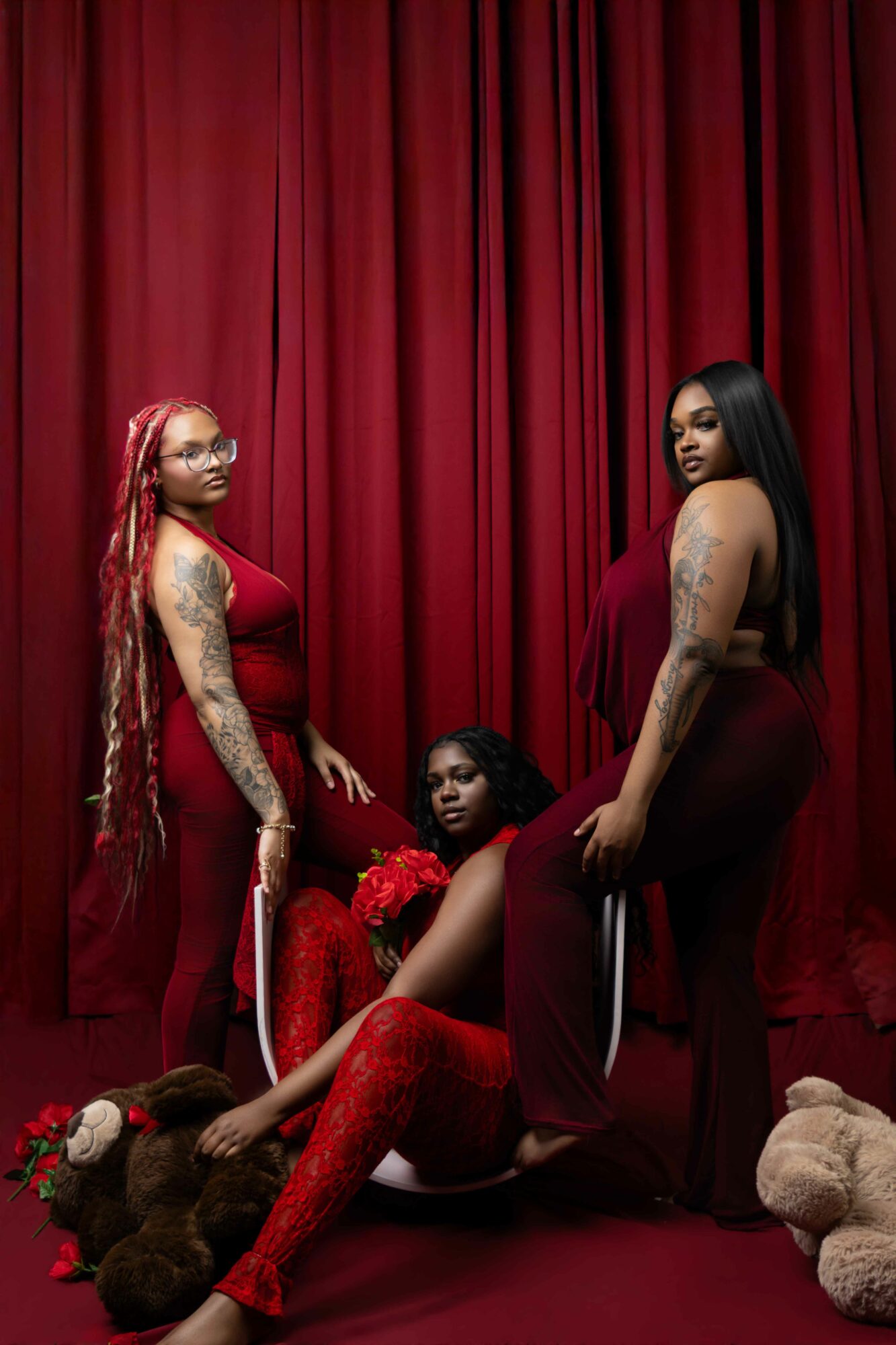 Three women in red outfits pose in front of a red curtain, with teddy bears on the floor, looking at the camera.