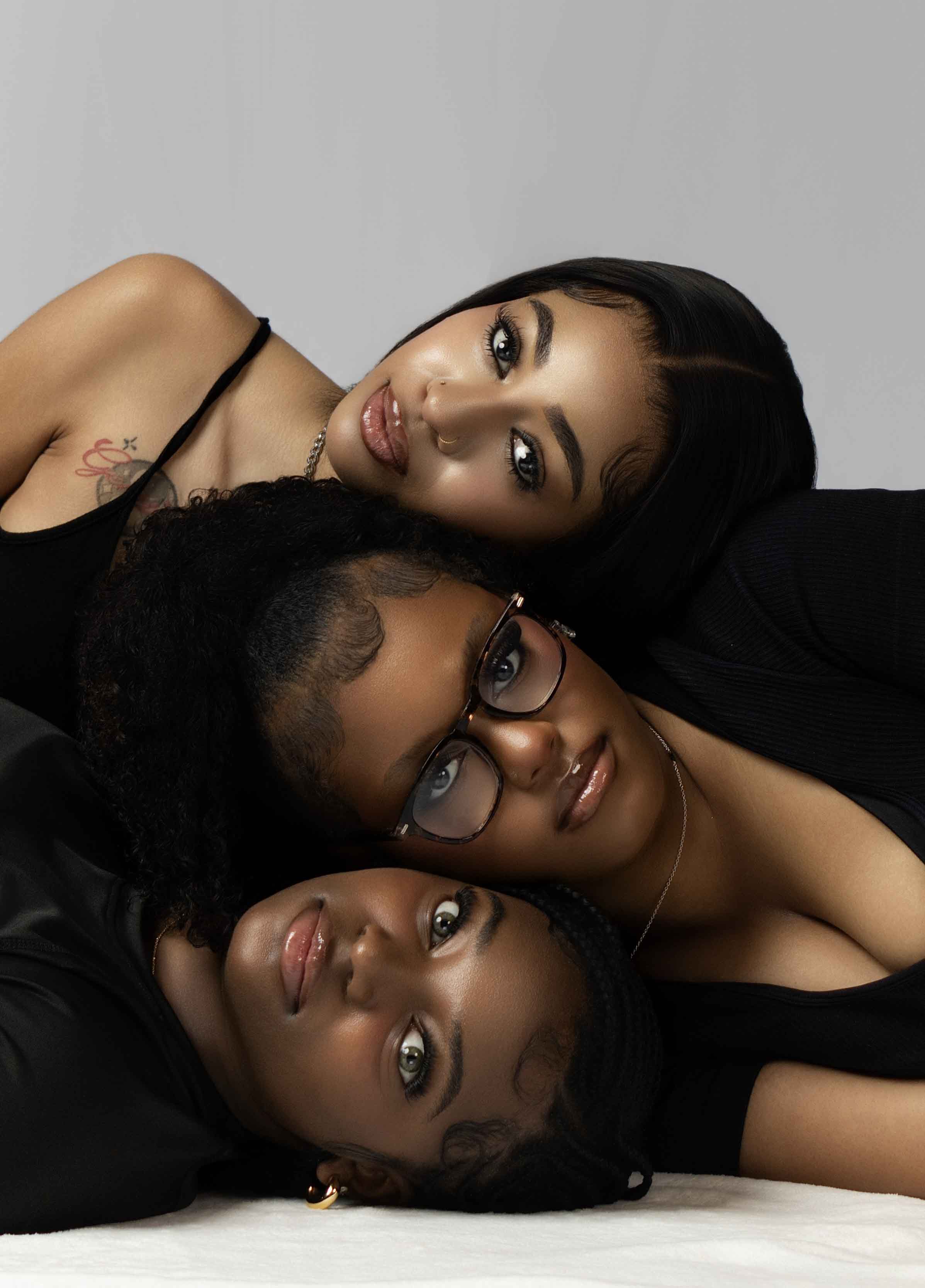 Three women lying close together, head to head, with neutral background, looking at camera, wearing black clothing.