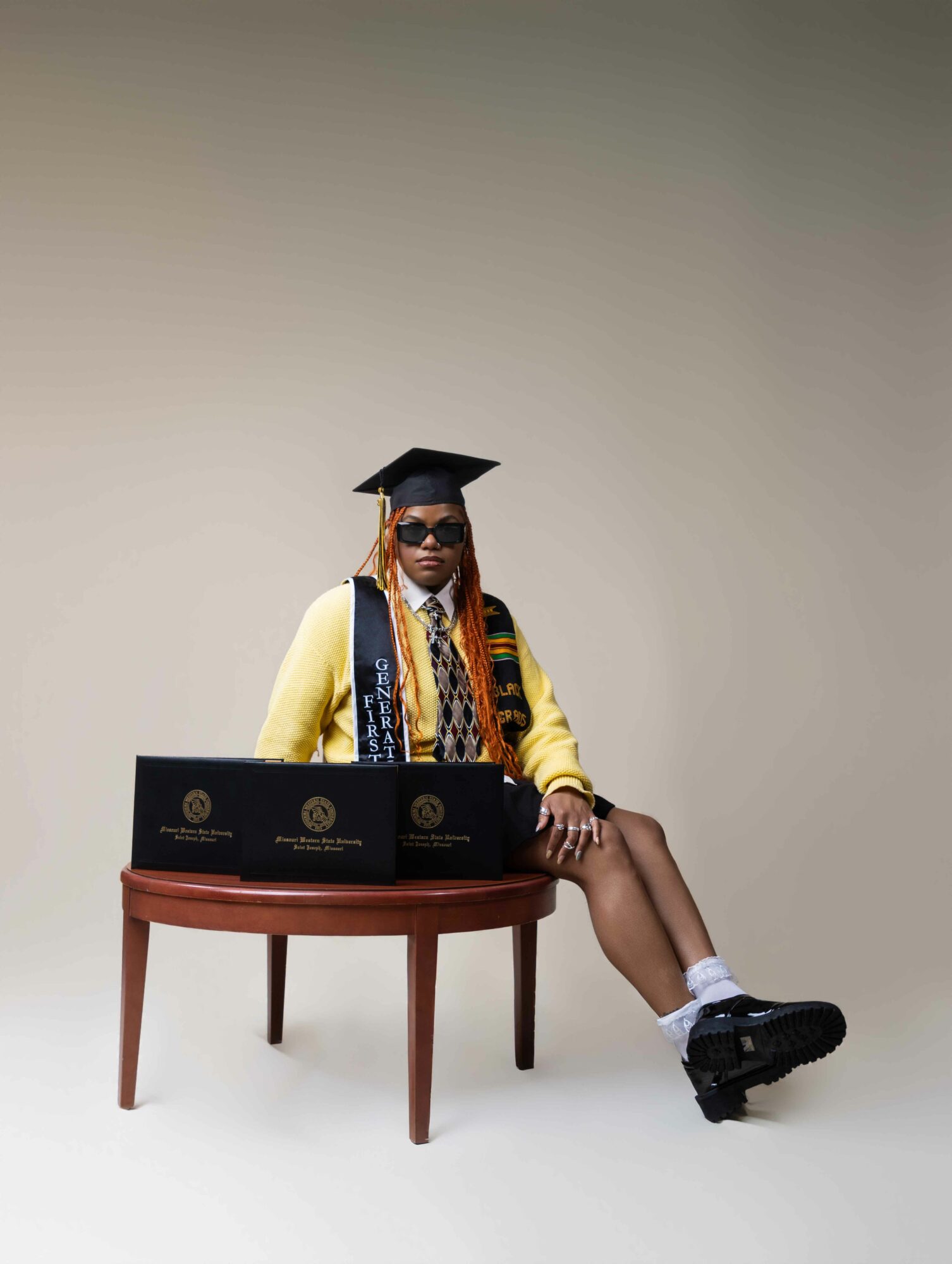 Person in graduation cap and gown sitting at a table with laptops, against a plain background.