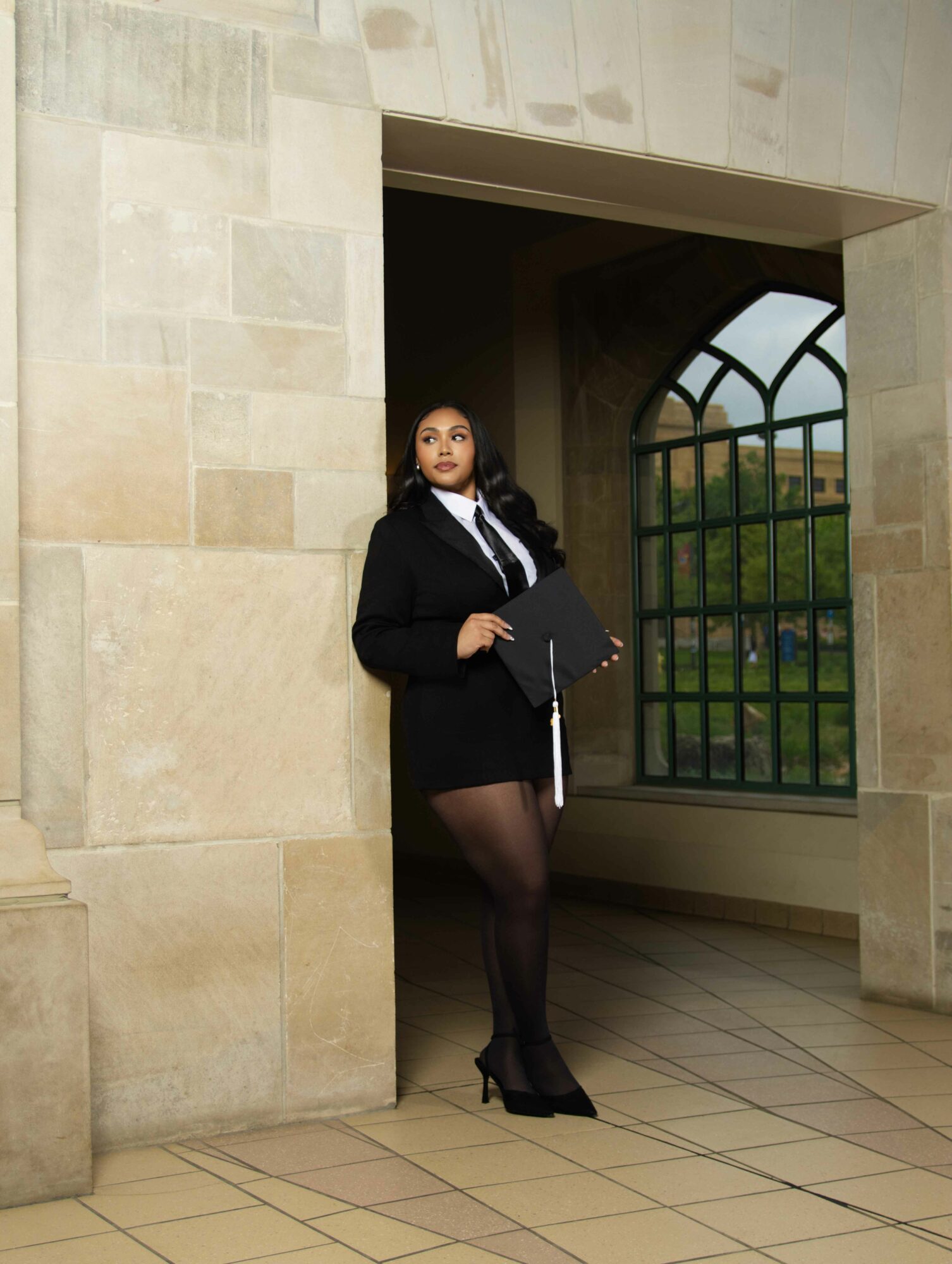 Woman in black blazer and skirt standing in an arched window nook holding a folder, looking to the side.