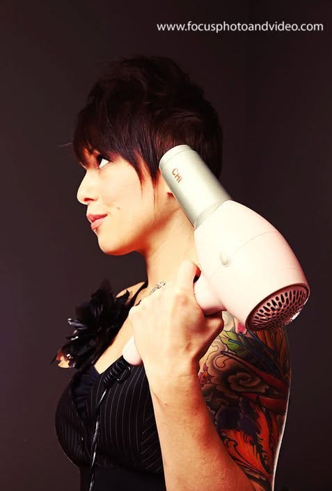 Woman with short hair holding a hairdryer on her shoulder, showing a colorful tattoo on her arm, against a dark background.