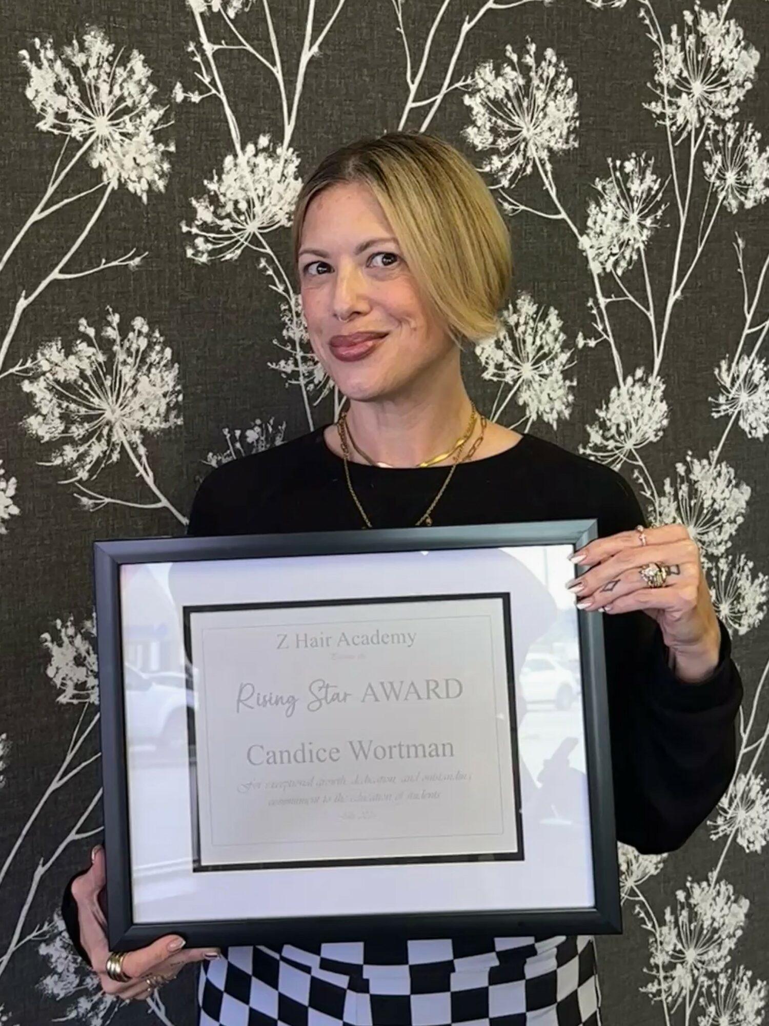 Woman holding framed award certificate, standing in front of floral patterned wallpaper, smiling, with short blonde hair, black top, and checkered skirt.
