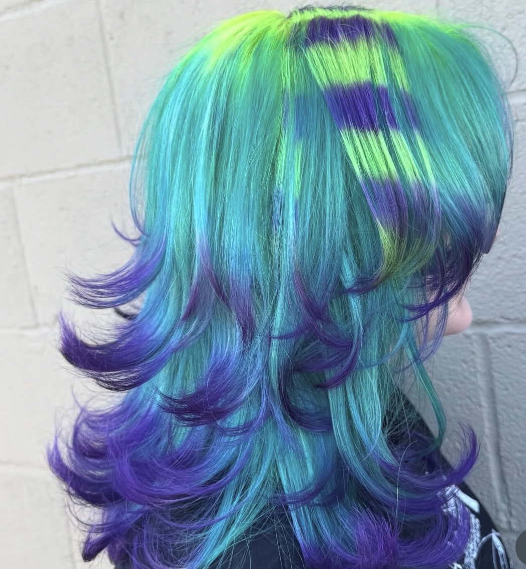 Side view of a person with vibrant, multi-colored hair in shades of green, blue, and purple, styled in loose curls.
