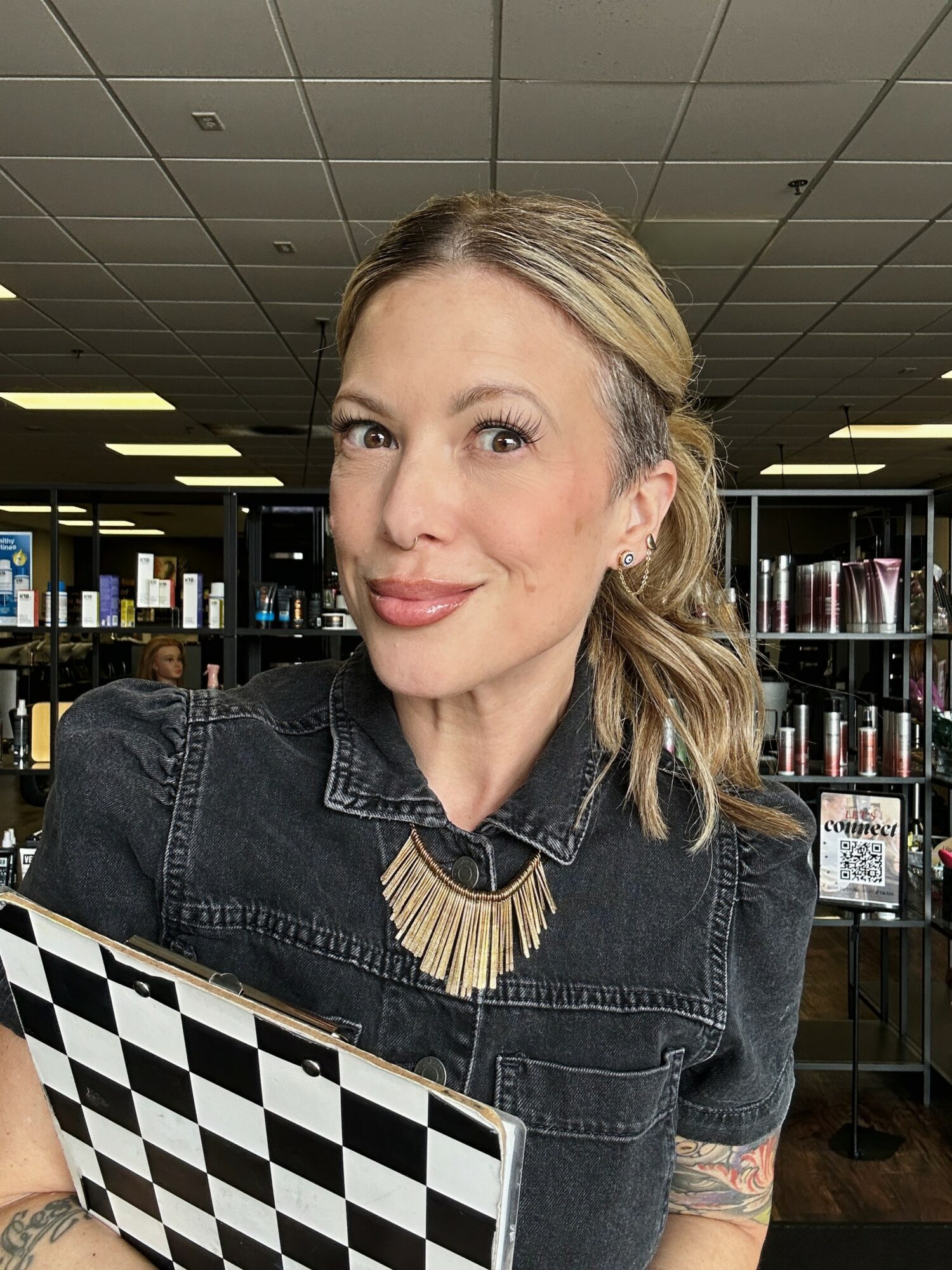Woman with blonde hair in a ponytail, wearing a black denim jacket and necklace, smiling in a store.