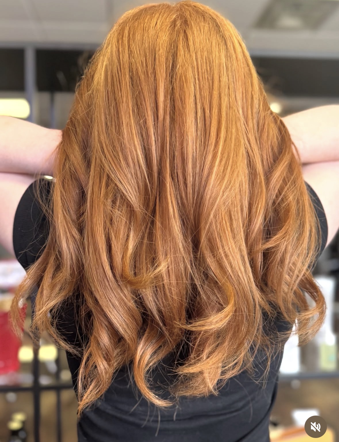 Back view of a person with long, wavy, reddish-orange hair, wearing a black top, in an indoor setting.