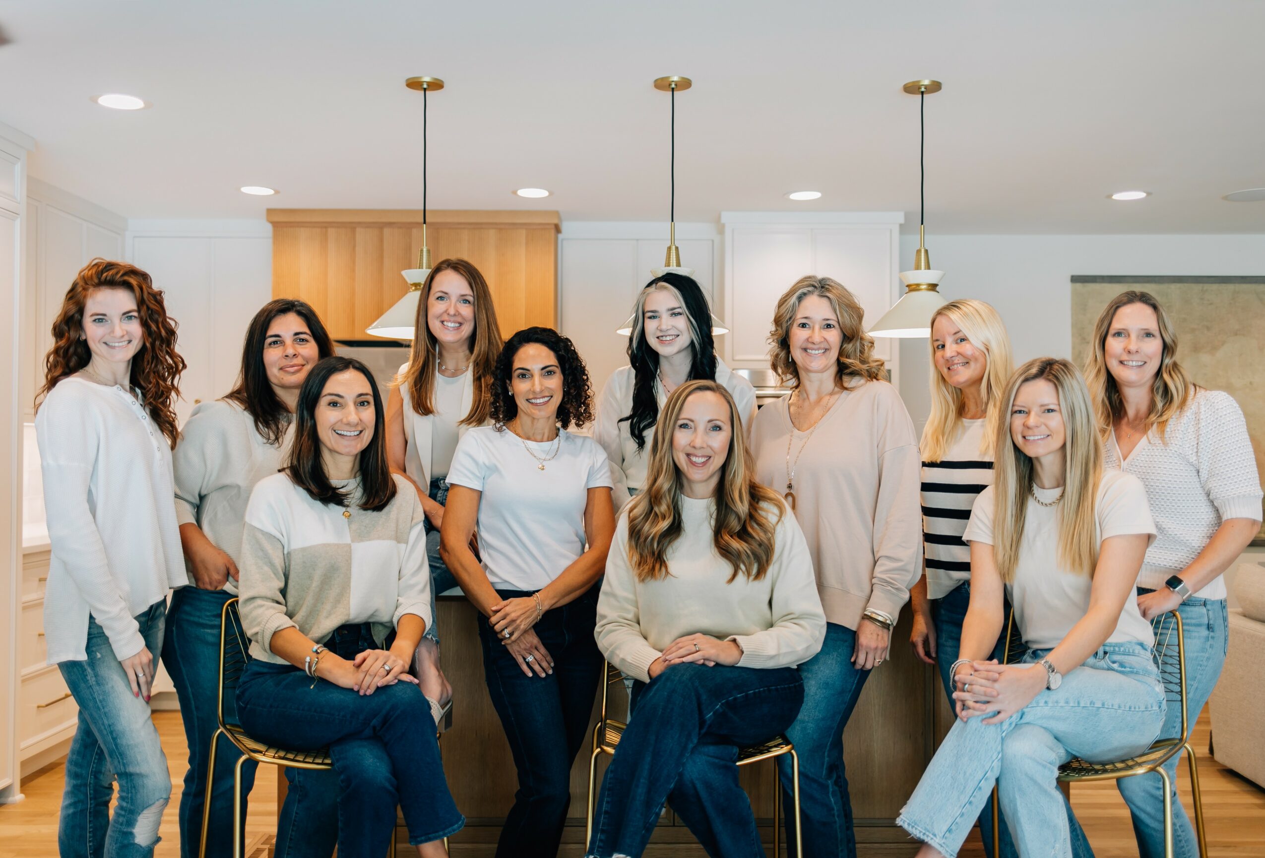 Group of eleven women standing and sitting in a modern kitchen, smiling, with pendant lights above.