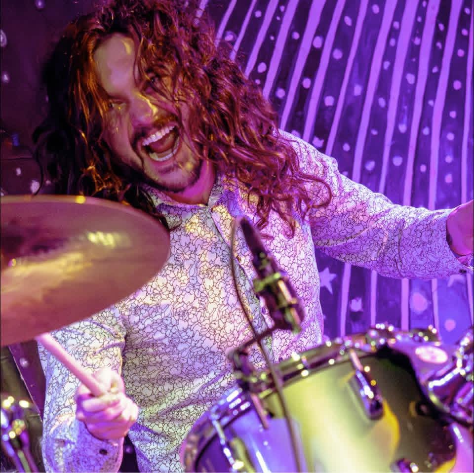 Person with curly hair playing drums, smiling, in a colorful stage setting.