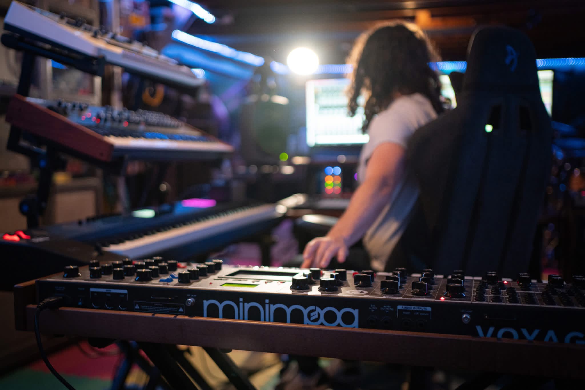 Person with curly hair working at music production equipment in a studio, illuminated by bright light, with keyboards and synthesizers around.