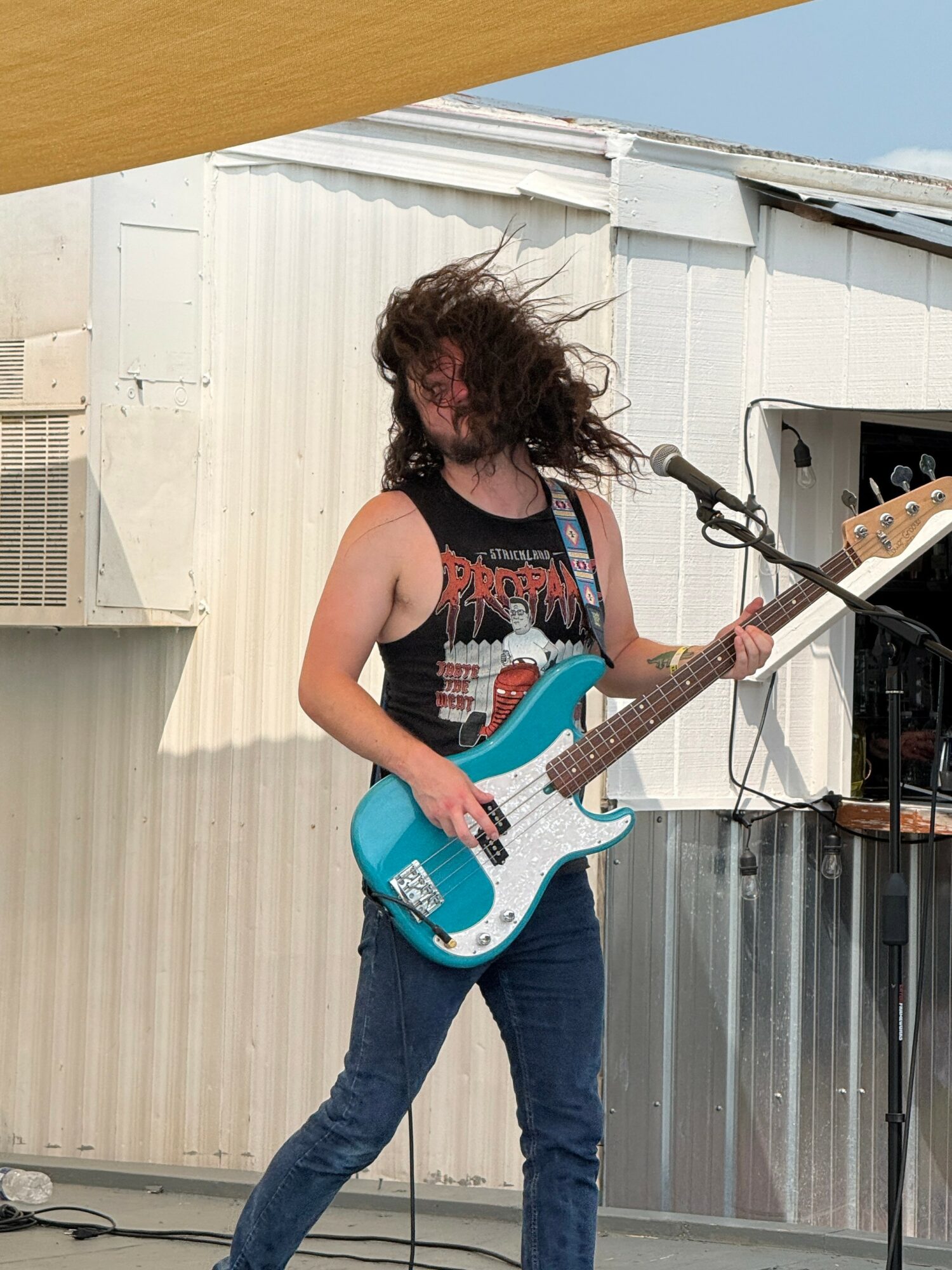 Person with long curly hair playing a blue electric guitar outdoors, standing near a microphone, with a metal building background.
