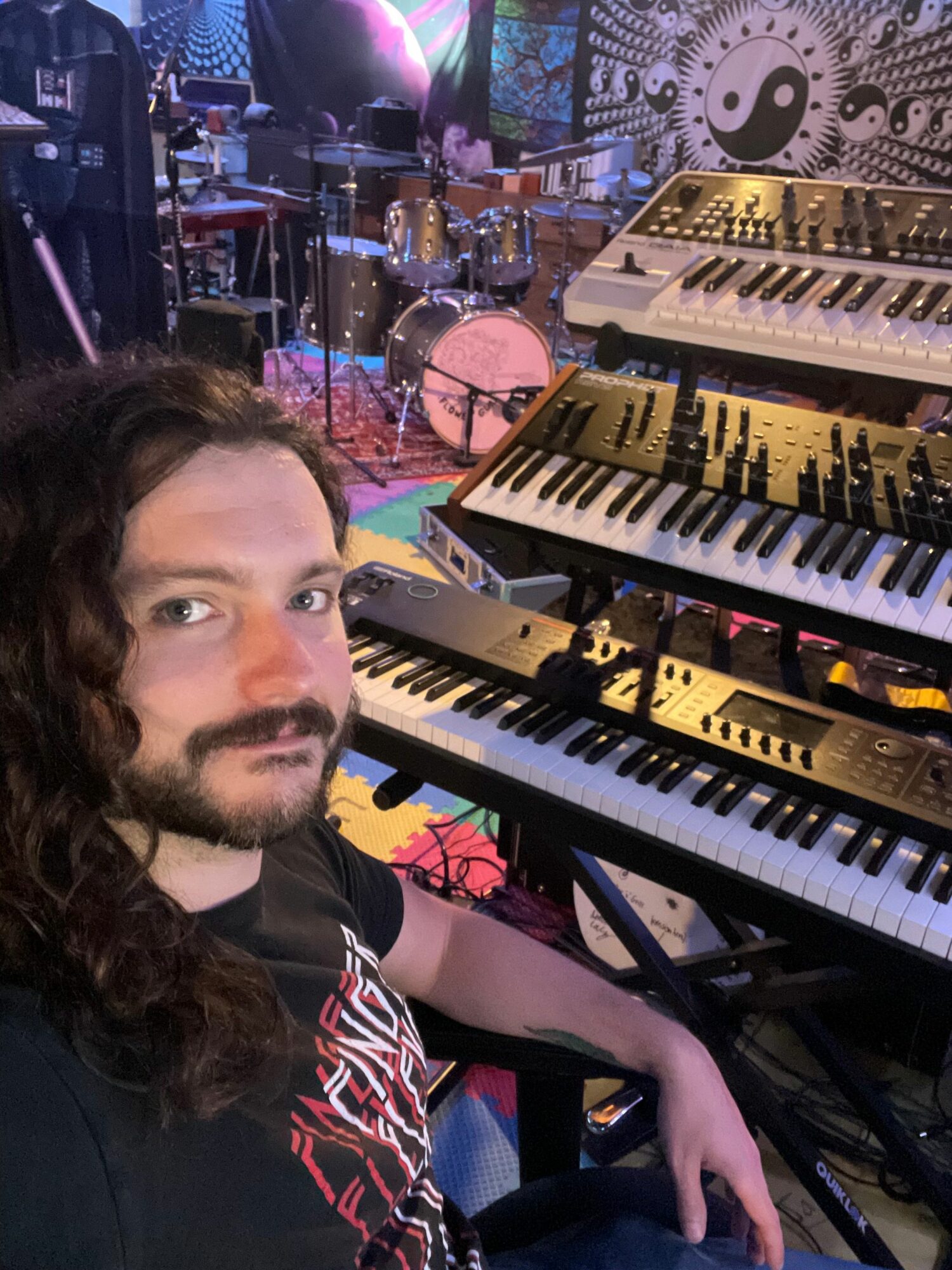 Person with long hair and beard lying near musical keyboards and equipment in a colorful setting.