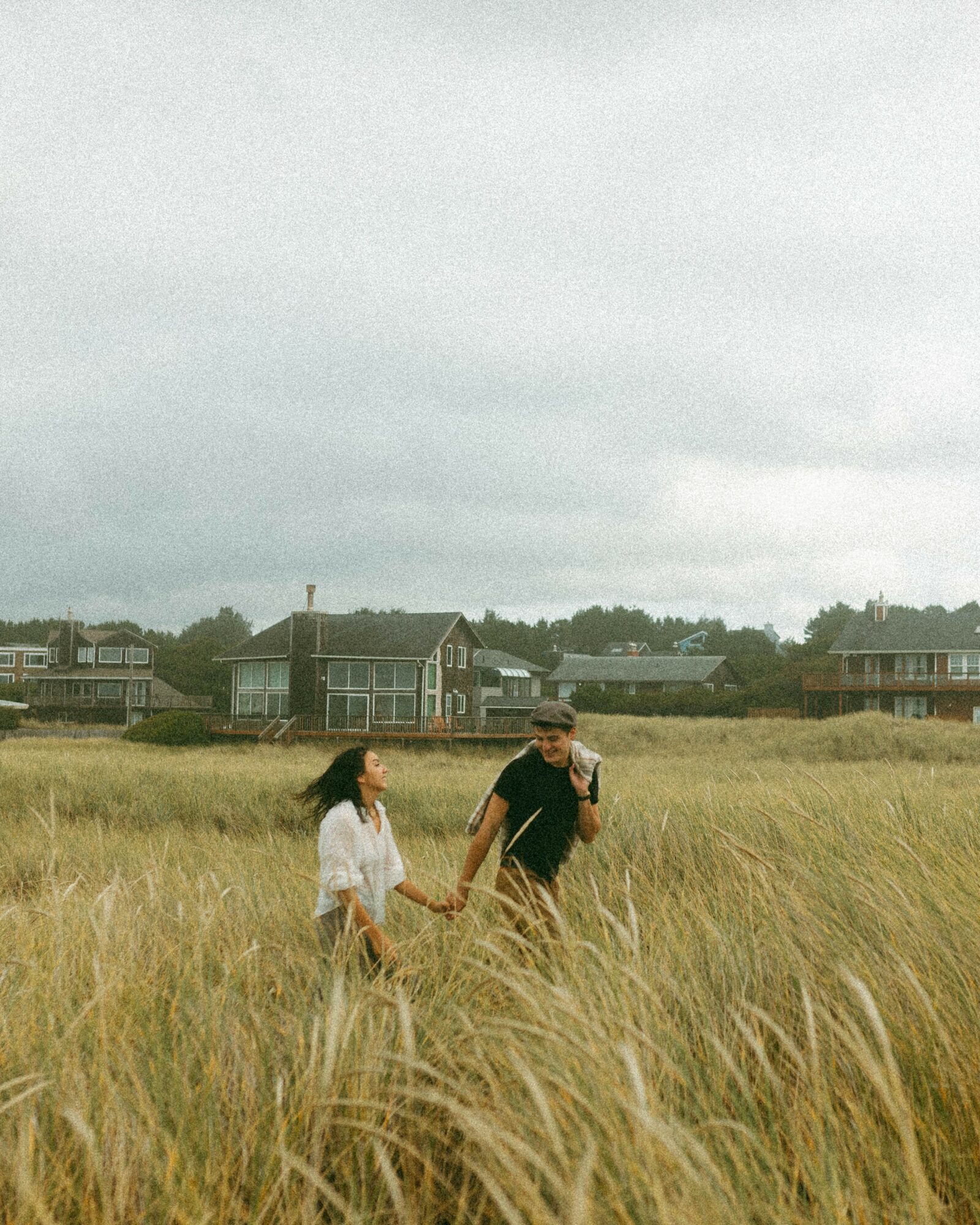 Two people holding hands walking through a grassy field under cloudy sky, with houses in the background.