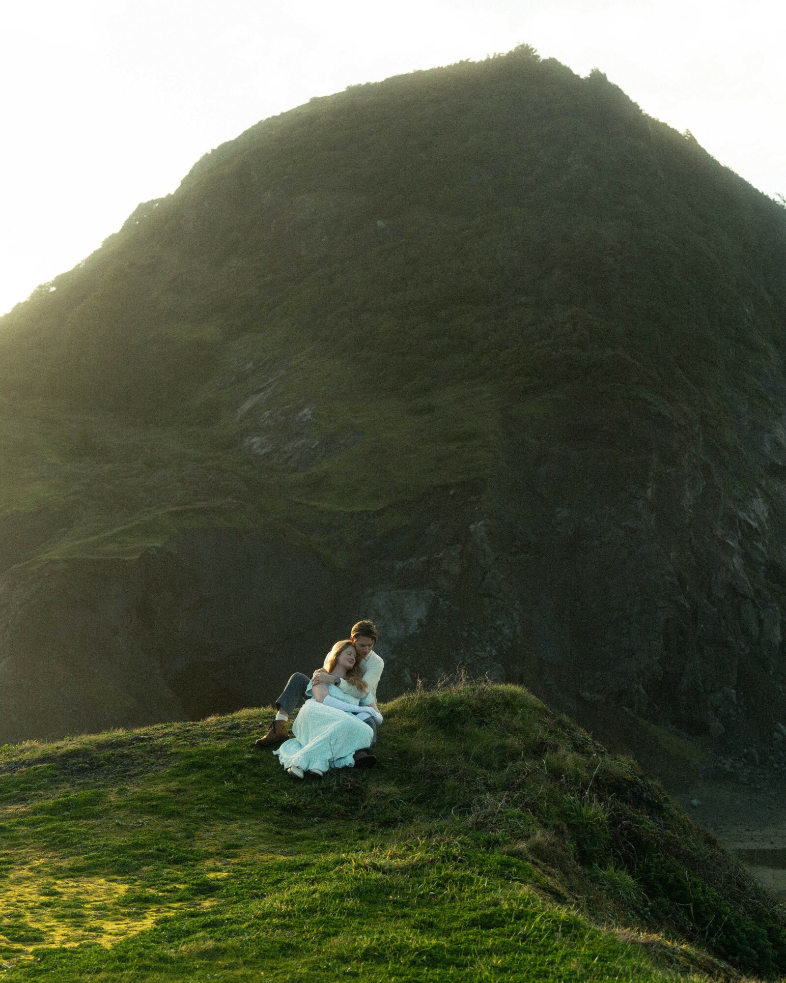 Person sitting on grass near a large mountain, with a backpack beside them, during daytime.