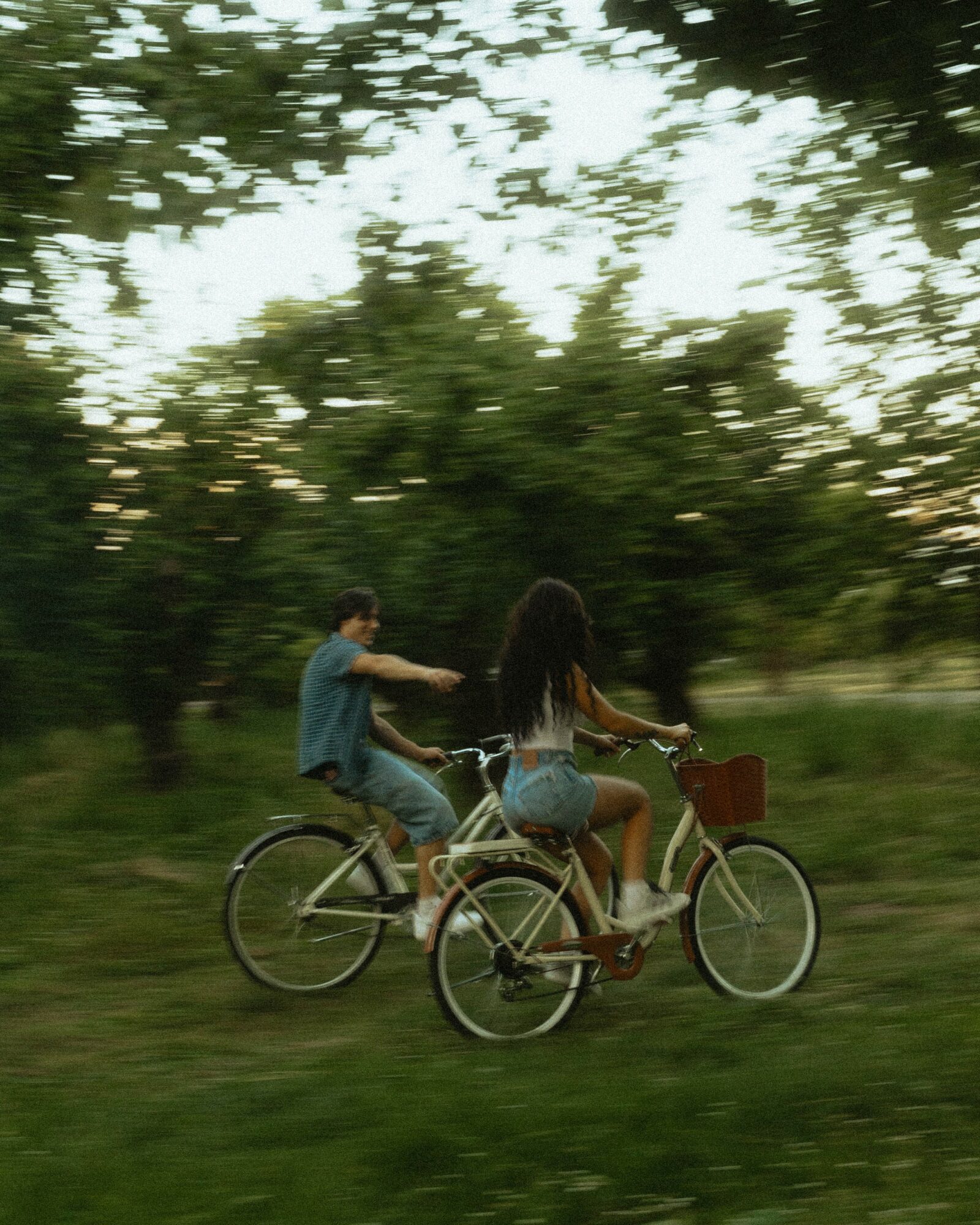 Two people riding bicycles on a grassy area with trees in the background, one person reaching out to the other.