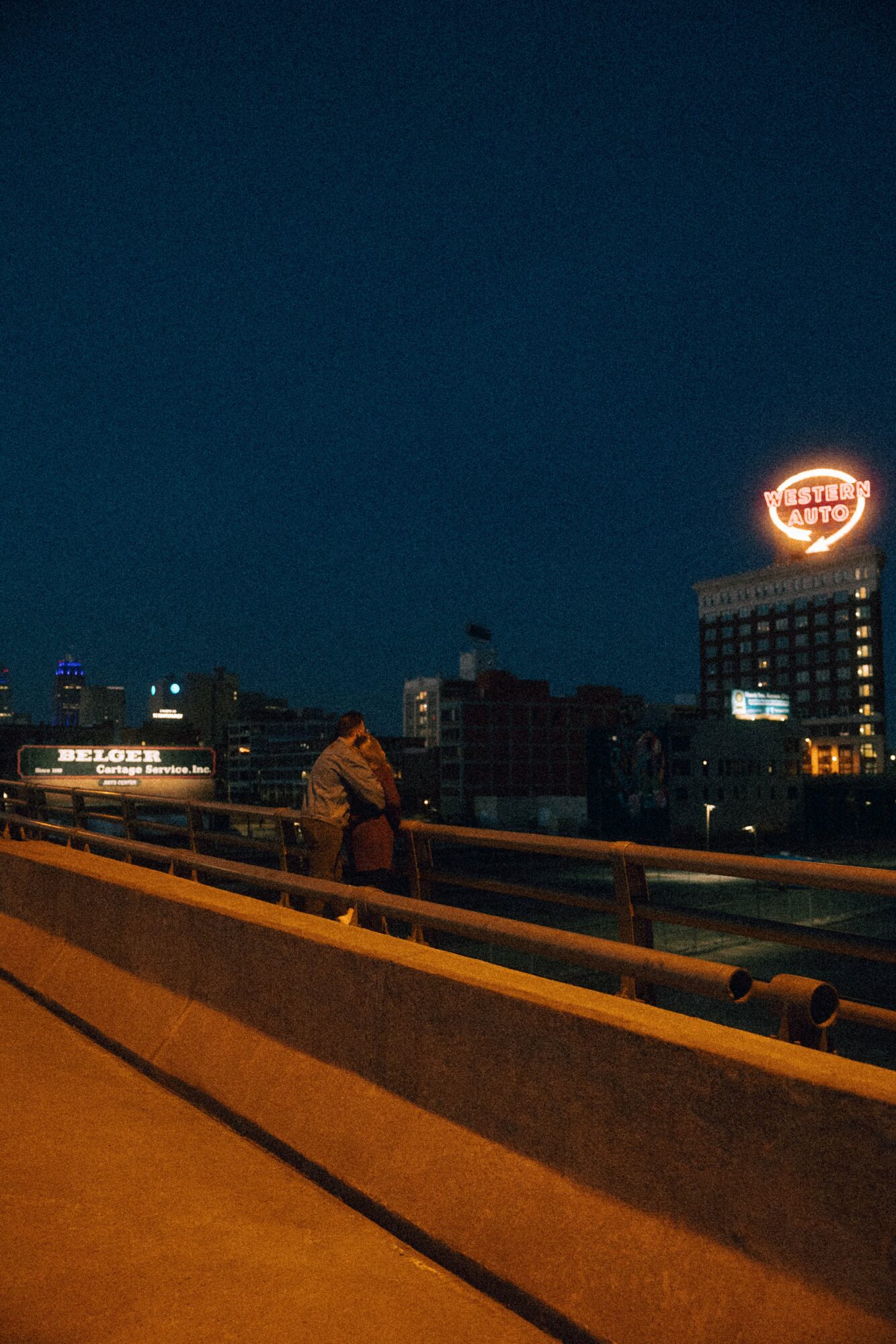 Person leaning on a railing on a bridge at night with city buildings and a lit sign in the background.