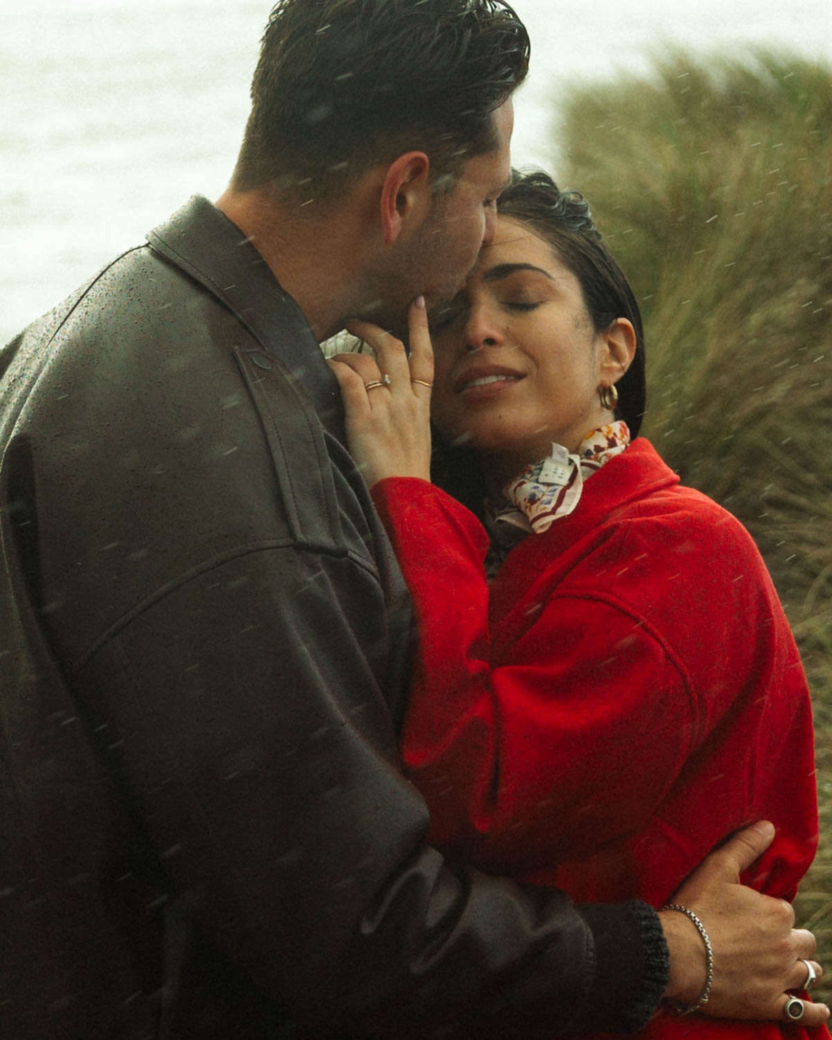A man and woman embrace outdoors near water, with the man gently touching the woman's face and the woman smiling.