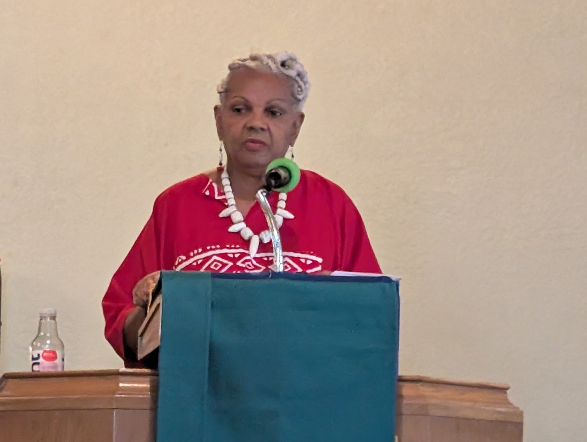 Woman with short gray hair speaking at a podium, wearing a red top and white necklace, with a microphone and water bottle nearby.
