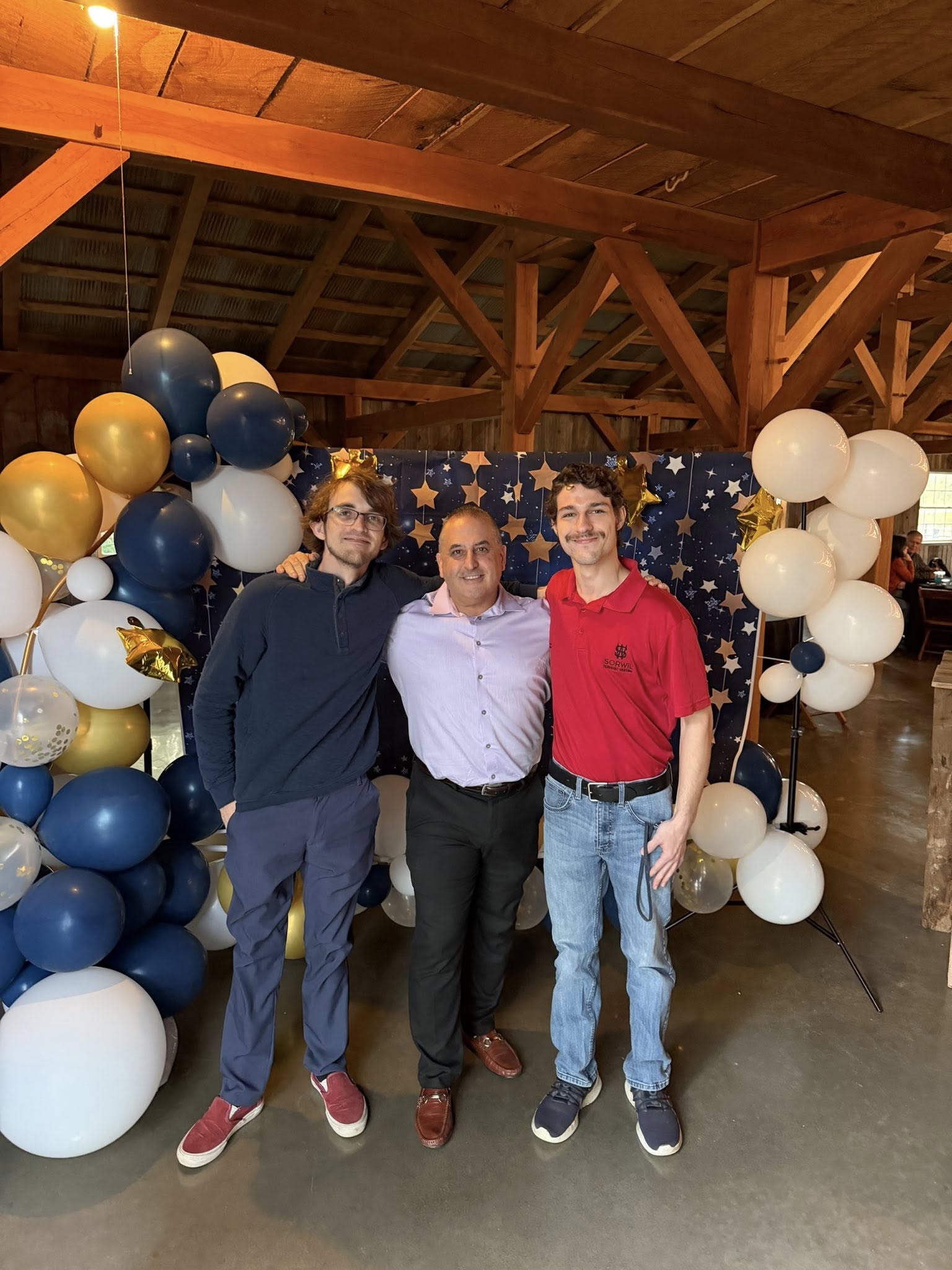 Three people standing together in front of a balloon decoration with blue, white, and gold balloons, inside a wooden structure.