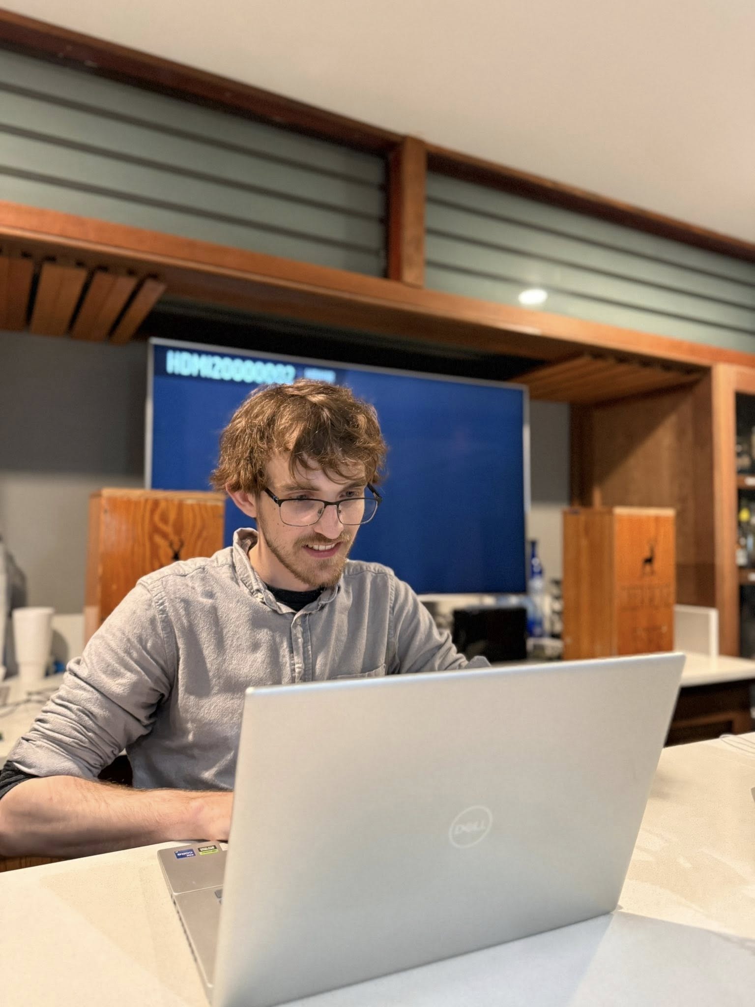 Young man with glasses and curly hair working on a laptop in a modern room with a large screen behind him.