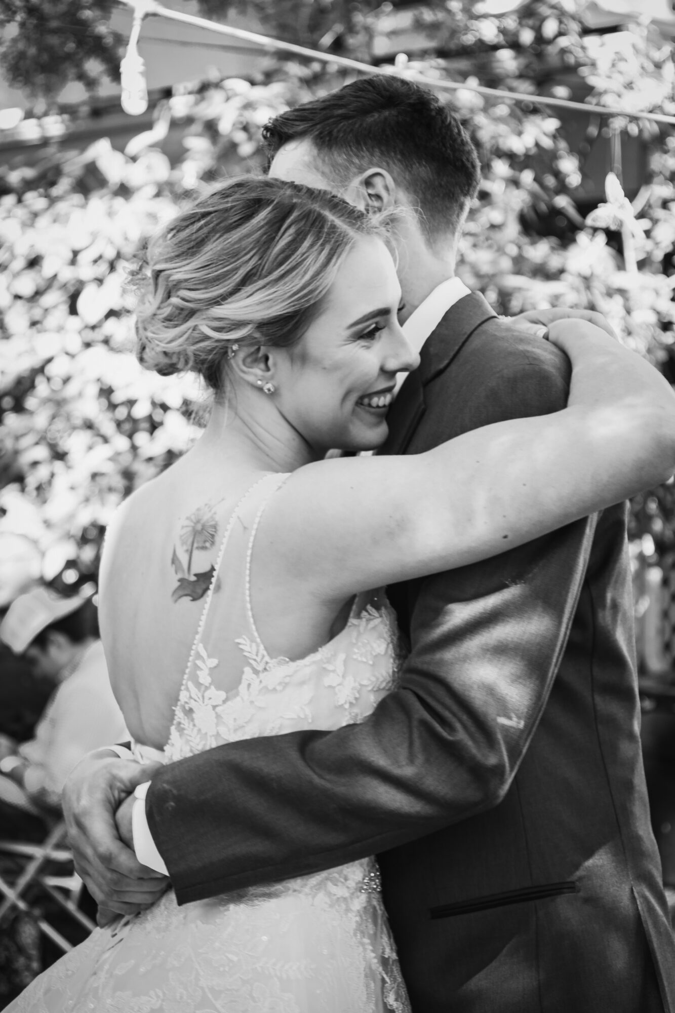A woman and man embrace outdoors, smiling, with trees and people in the background.