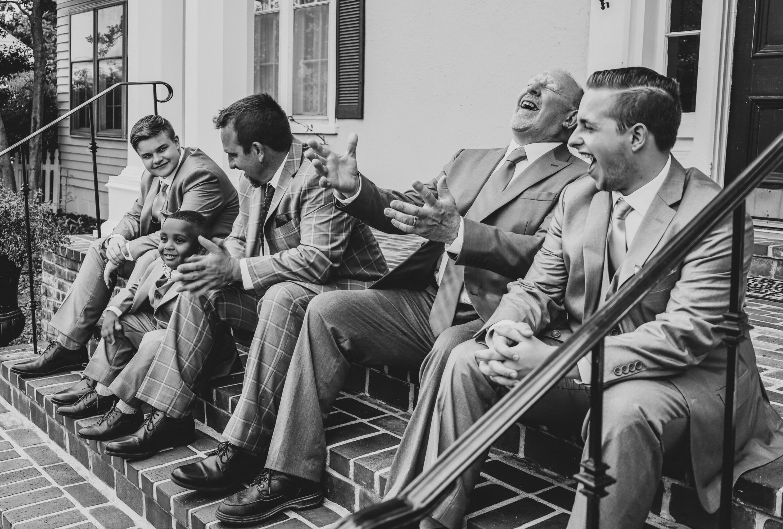 Six men and boys sit on steps outside a building, laughing and talking together, dressed in suits and casual clothing.