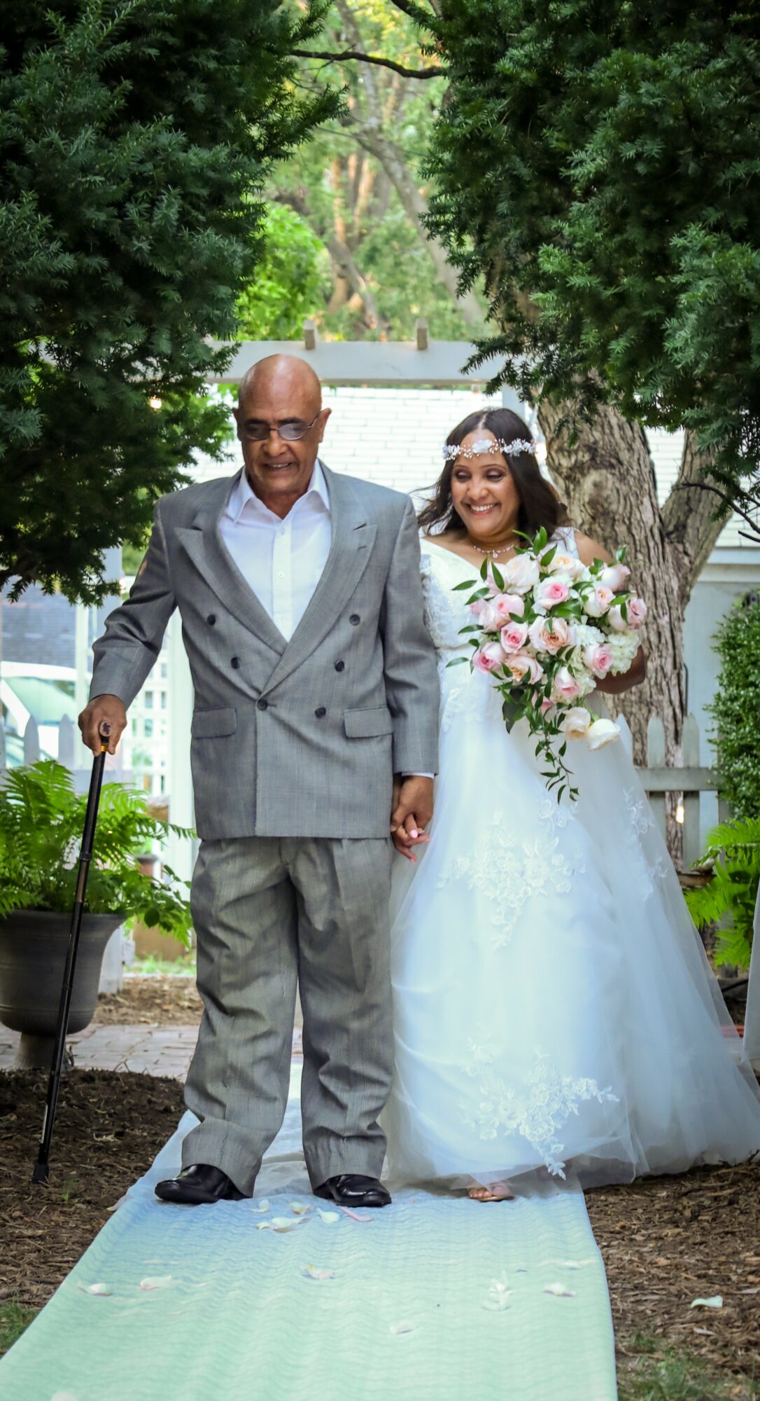 A bride in a white wedding dress holding a bouquet, walking with a man in a gray suit outdoors.