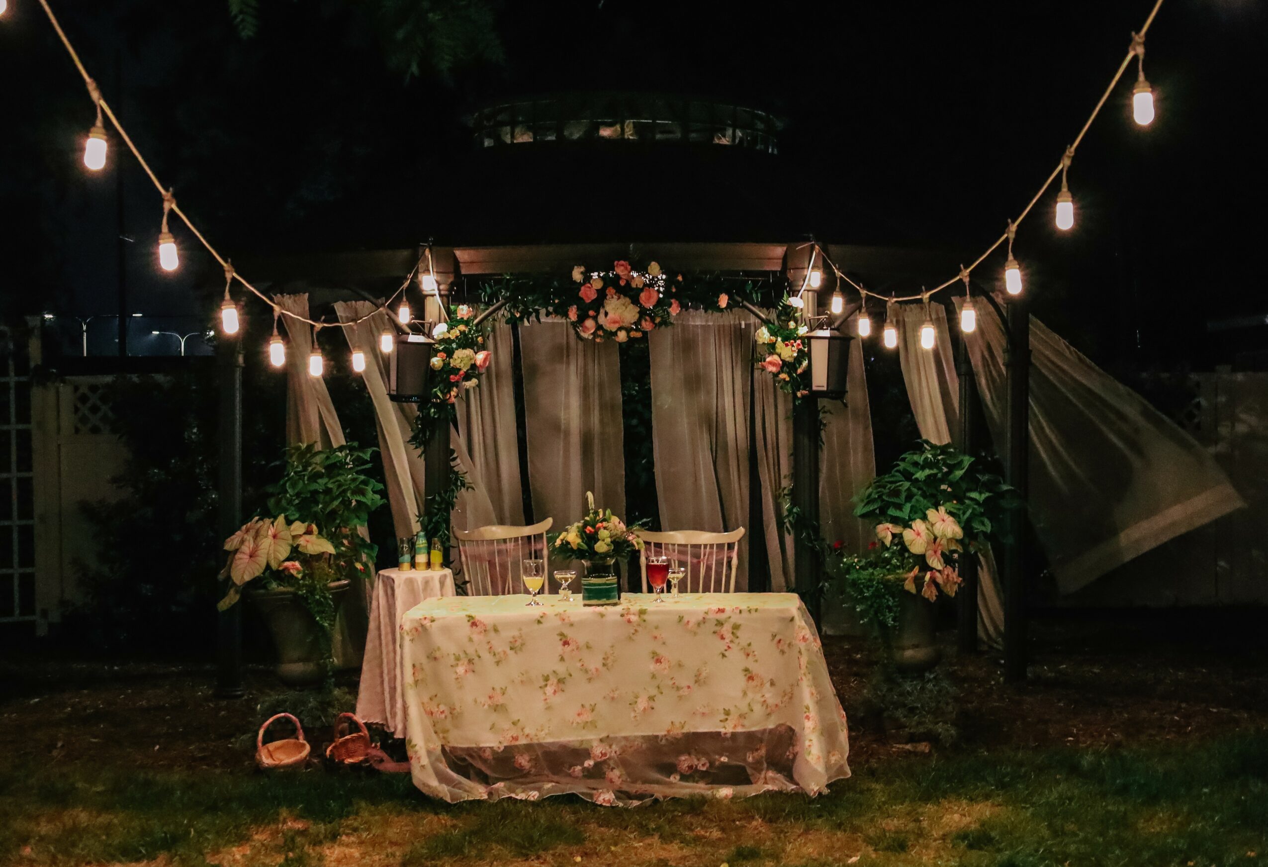 Nighttime outdoor scene with a decorated table, string lights, floral arrangements, and a backdrop, illuminated softly.