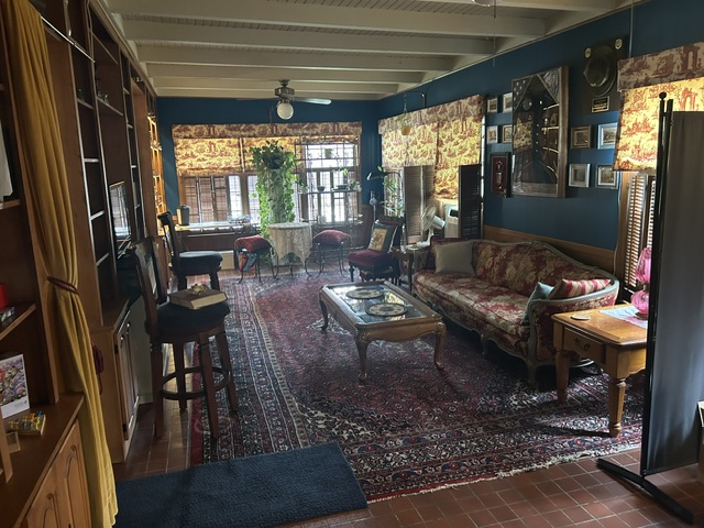 Living room with large windows, patterned rug, sofa, armchairs, and wooden furniture, decorated with photos and plants.