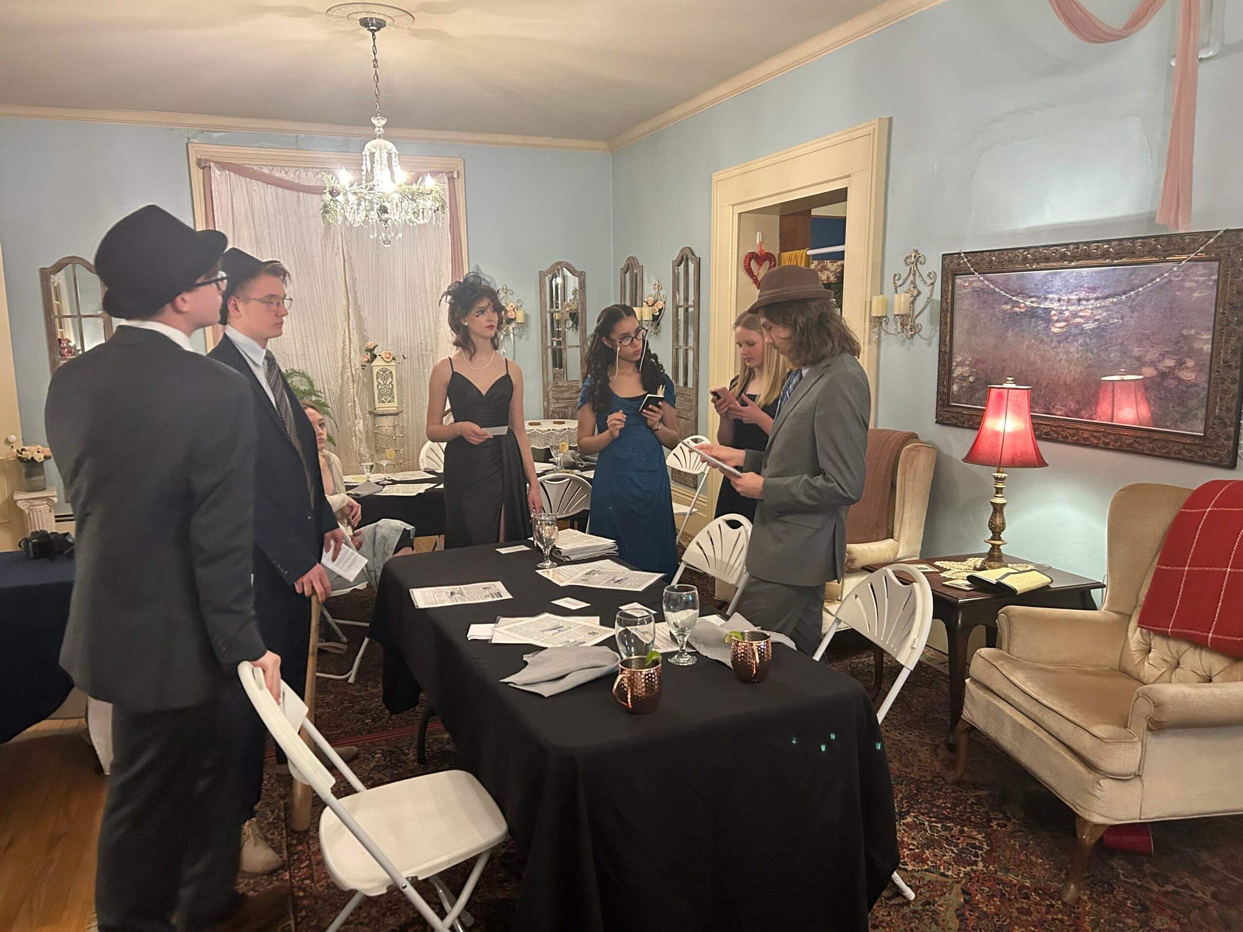 People dressed in vintage clothing gather around a table in a decorated room with a chandelier, sofa, and television.