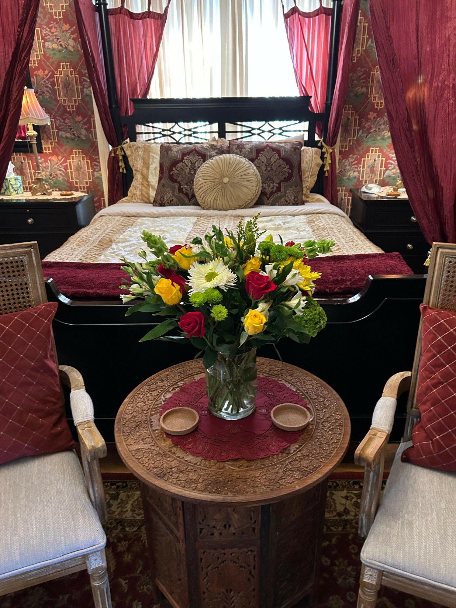 A dining room with a table holding a colorful flower arrangement, surrounded by chairs, with curtains and patterned wallpaper in the background.