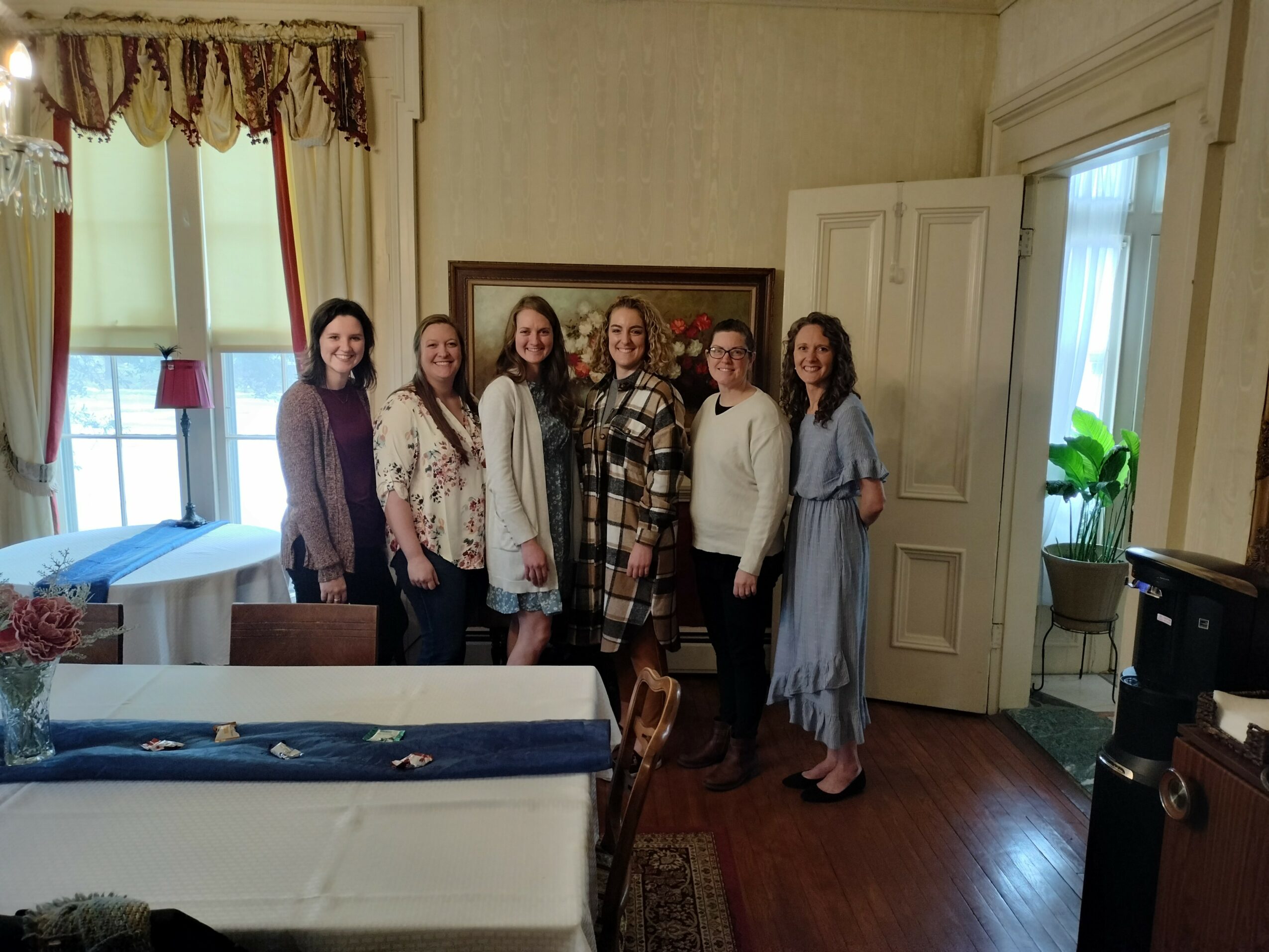 Seven women stand together in a room with a window, curtains, and a door, smiling at the camera.