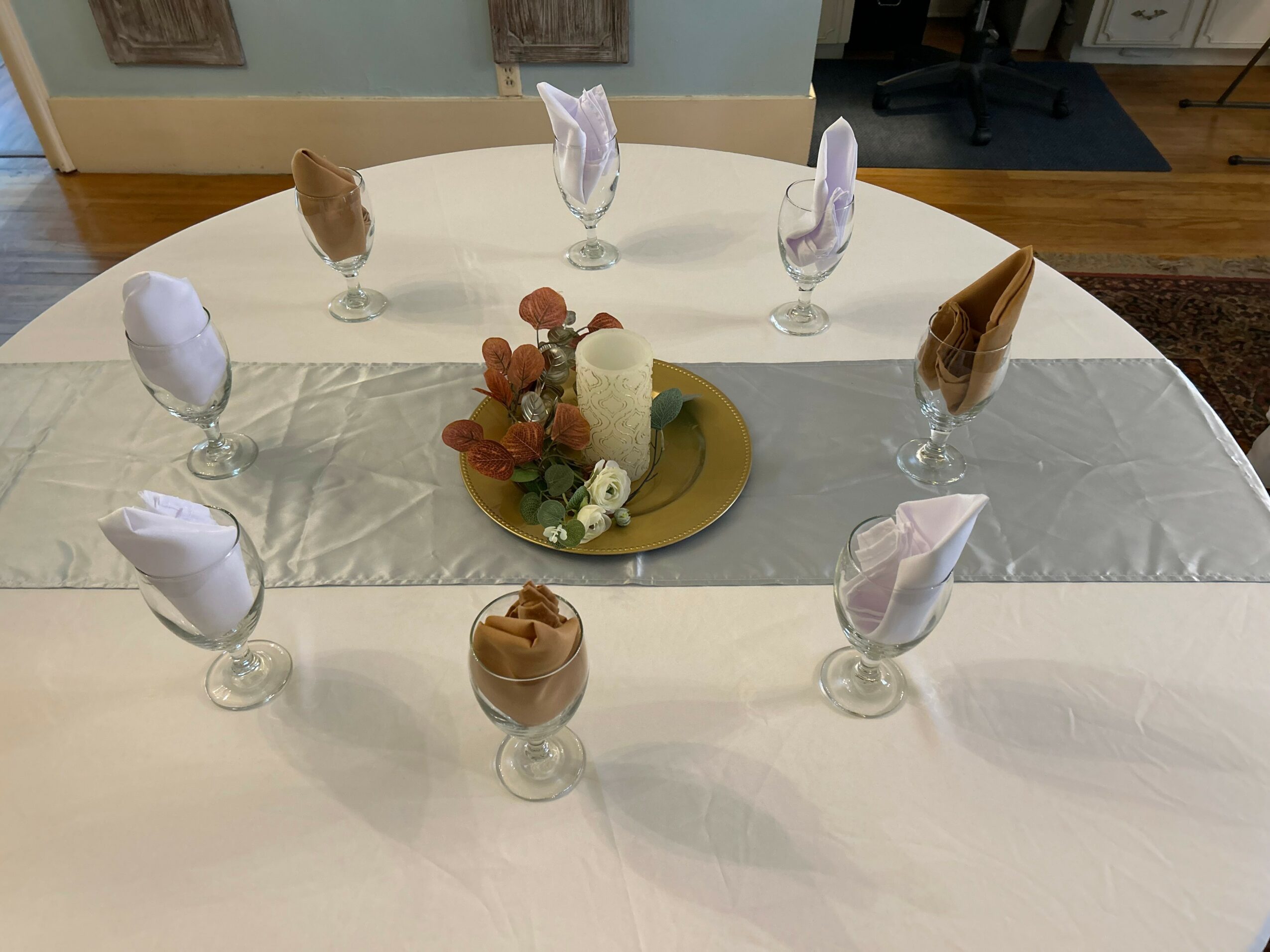 Round table with eight glasses, each with a folded napkin, and a centerpiece with flowers and a candle.