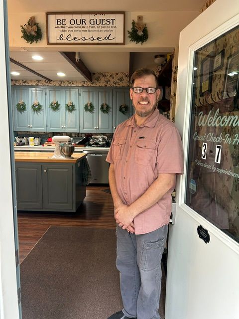 Man with glasses and a pink shirt standing in a kitchen with a sign that reads 'Be Our Guest' behind him.