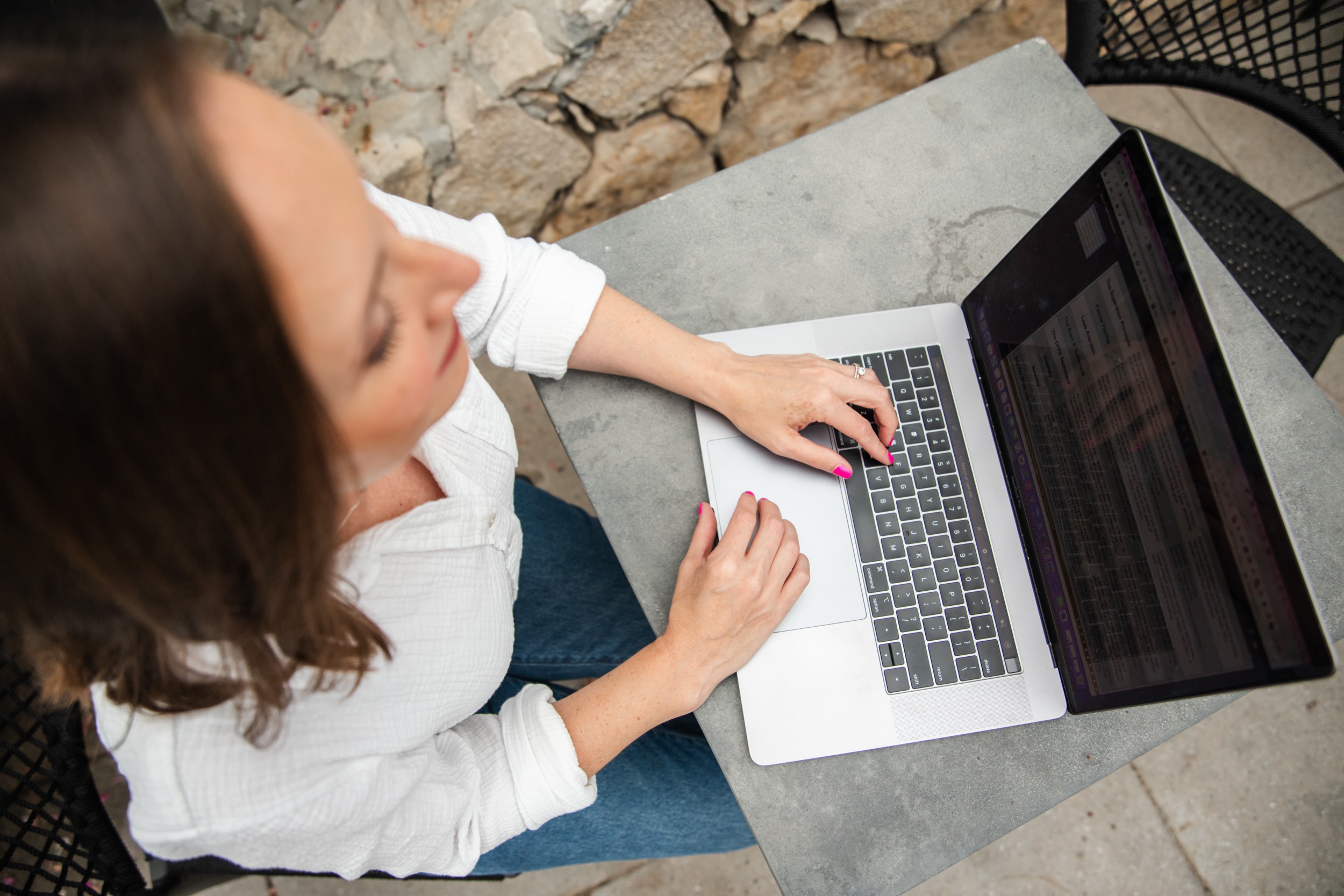 Woman using a laptop outdoors at a table, with stone wall background, viewed from above.