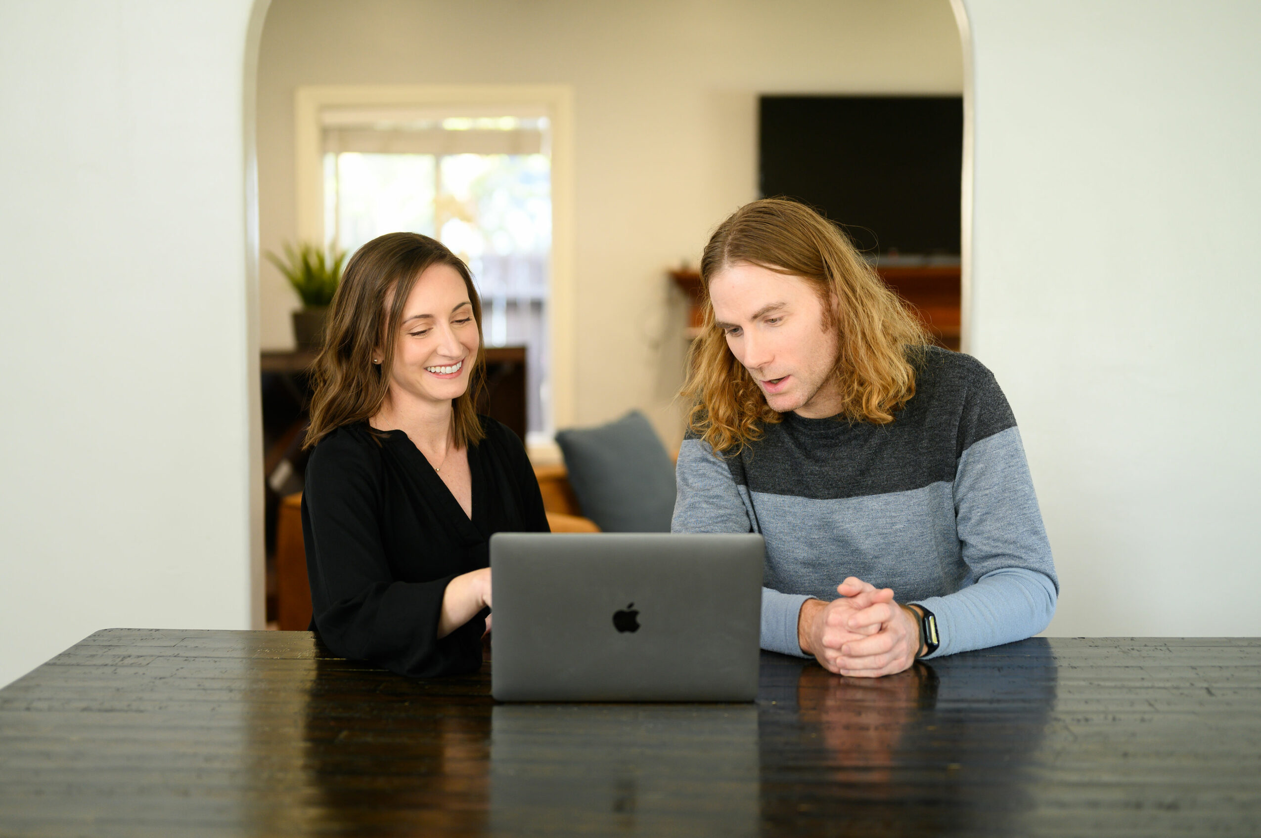 Two women sit at a table looking at a laptop, smiling and engaged in conversation, in a home setting.