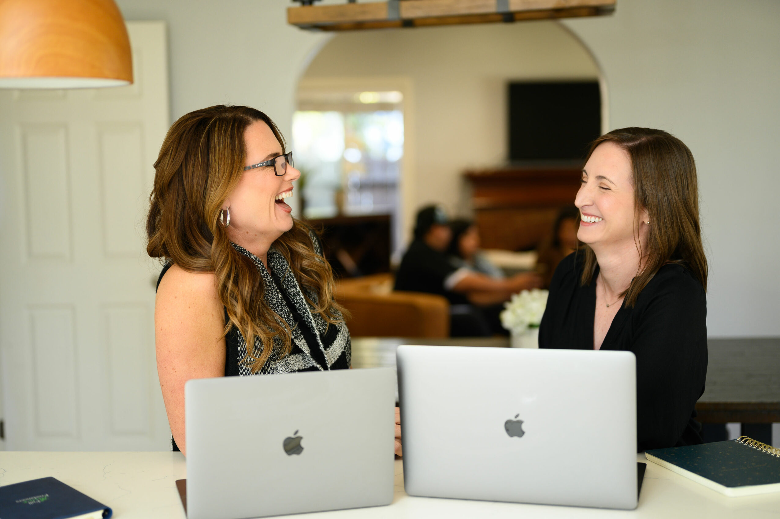 Two women laughing and talking at a table with laptops in a bright room.