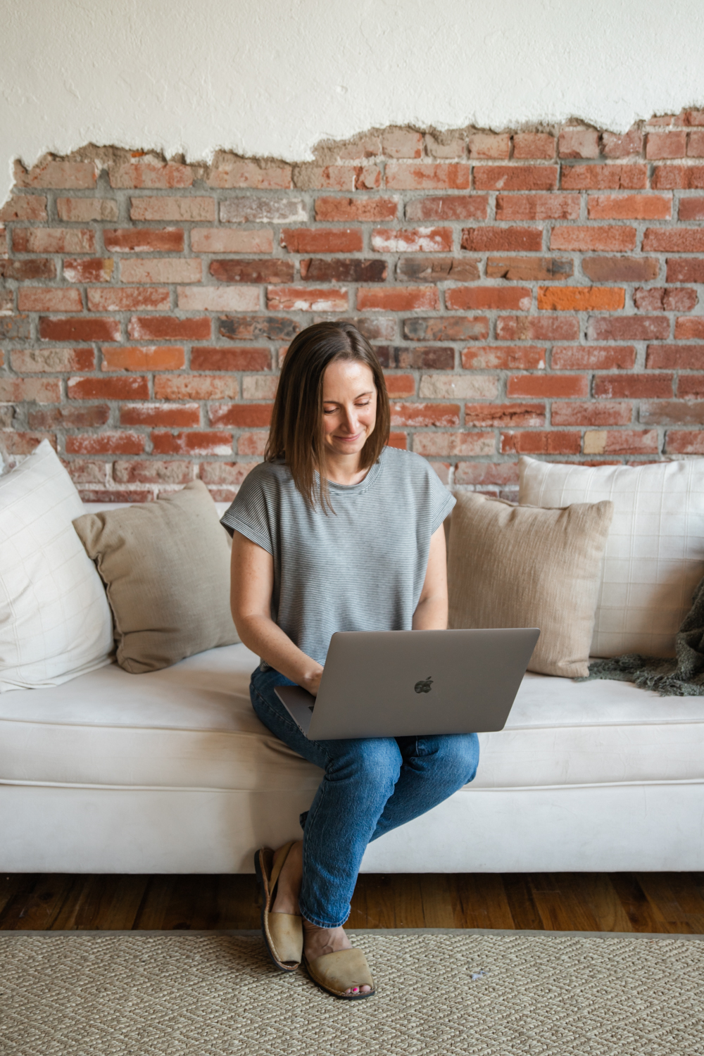 Woman sitting on a sofa using a laptop, brick wall background, casual clothing, smiling.