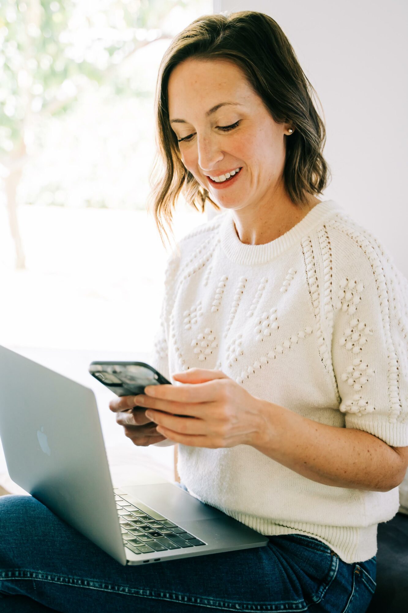 Woman smiling while looking at her phone, sitting with a laptop on her lap, in a bright room.