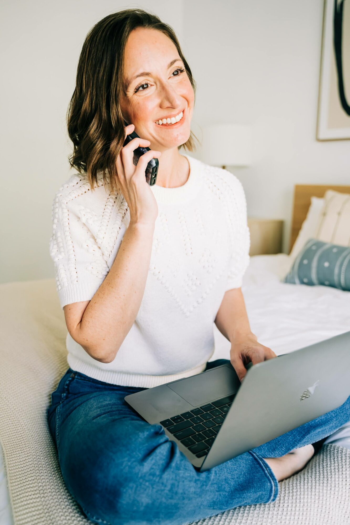 Woman smiling, talking on phone, using laptop in a bedroom, wearing a white top and jeans.