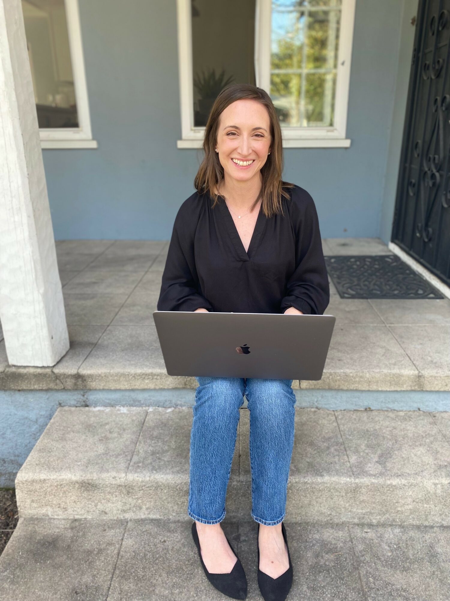 Woman sitting on steps with laptop, smiling, in front of a blue house with windows and a door.