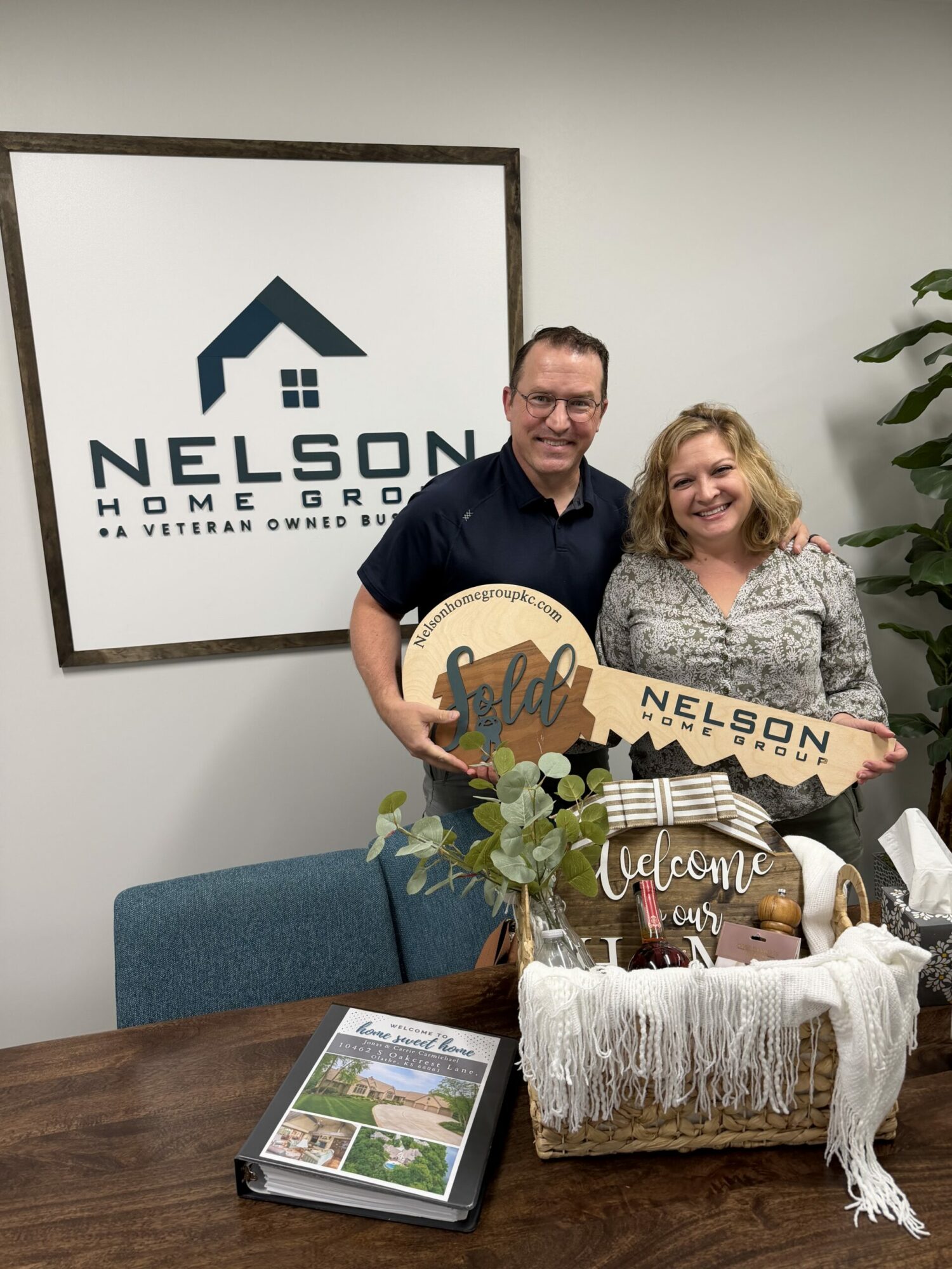 Two people standing behind a table with a welcome sign and a gift basket, smiling, in an office setting.