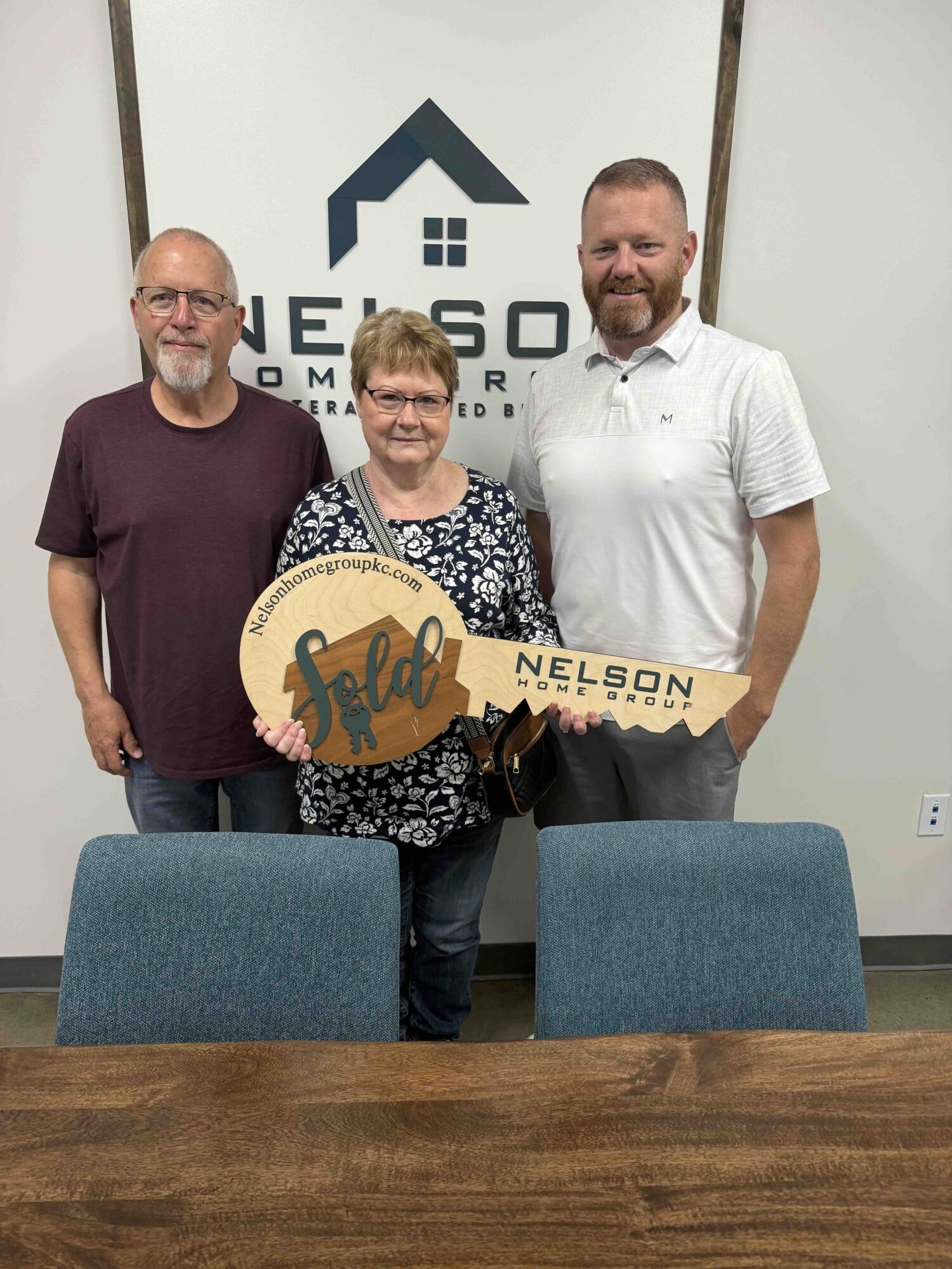 Three people standing indoors, holding a large key-shaped sign, in front of a logo on the wall.