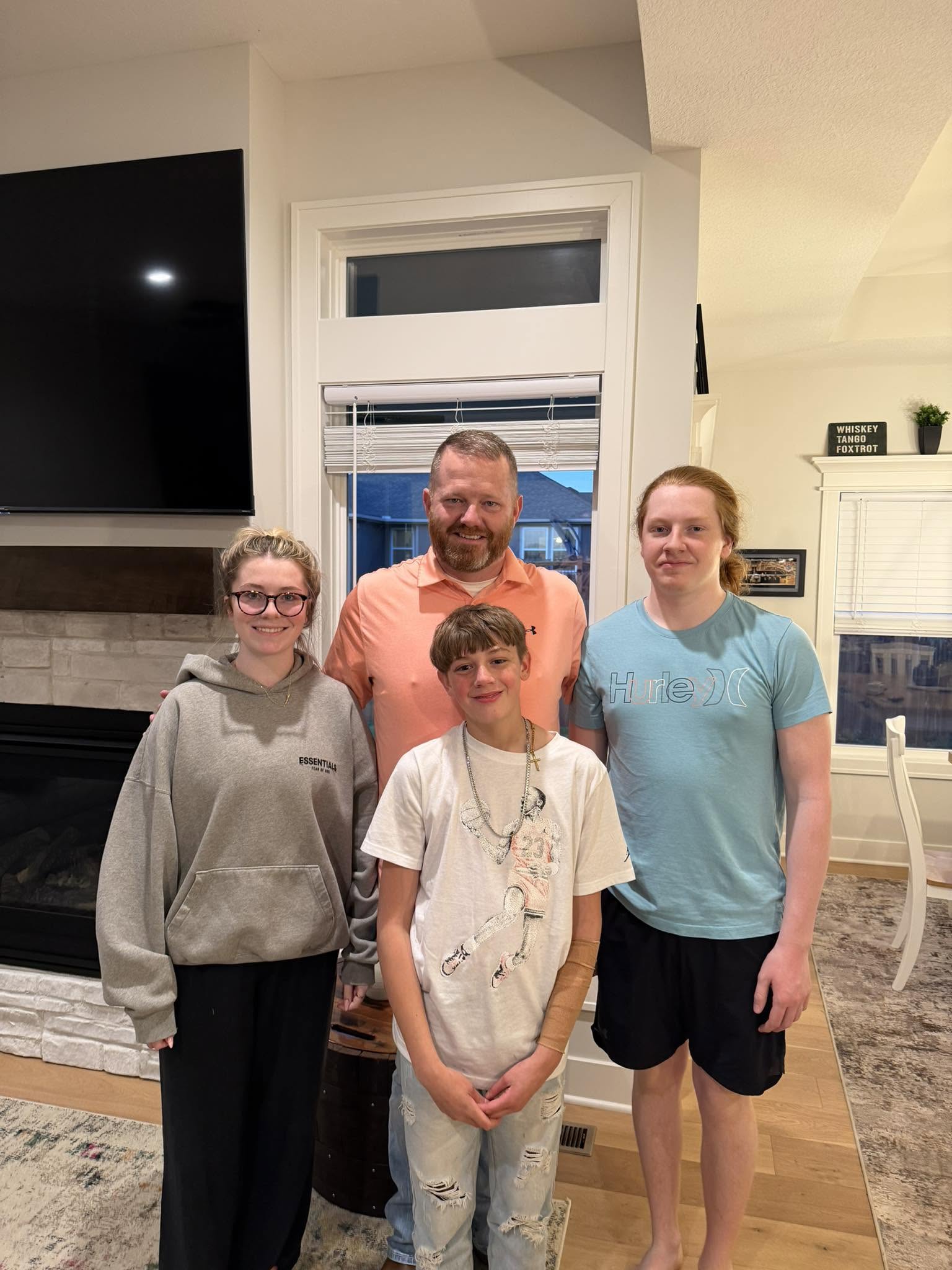 Four people standing indoors in front of a window, smiling, with a television and fireplace in the background.