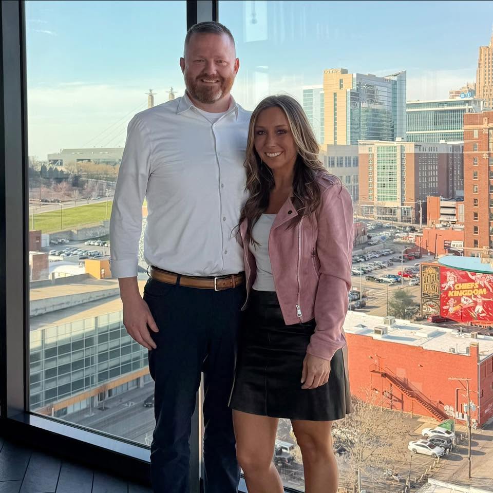 A man and woman stand together indoors with a cityscape view behind them, smiling at the camera.
