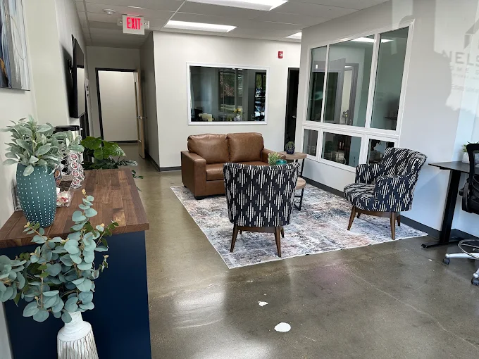 Waiting room with a brown sofa, two patterned chairs, a rug, and a windowed wall, with a wooden table and plants nearby.