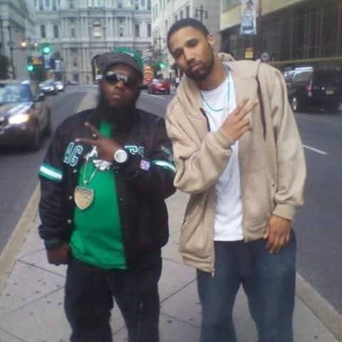 Two men standing on a city street, one wearing sunglasses and a black jacket, the other in a beige jacket, both making hand gestures.