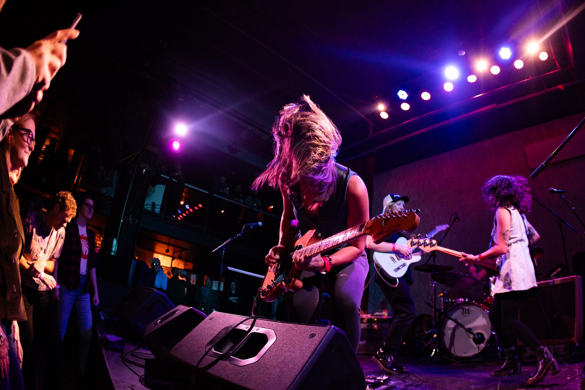 Musicians perform on stage with colorful lights, one playing guitar with long hair, others in the background, audience visible.